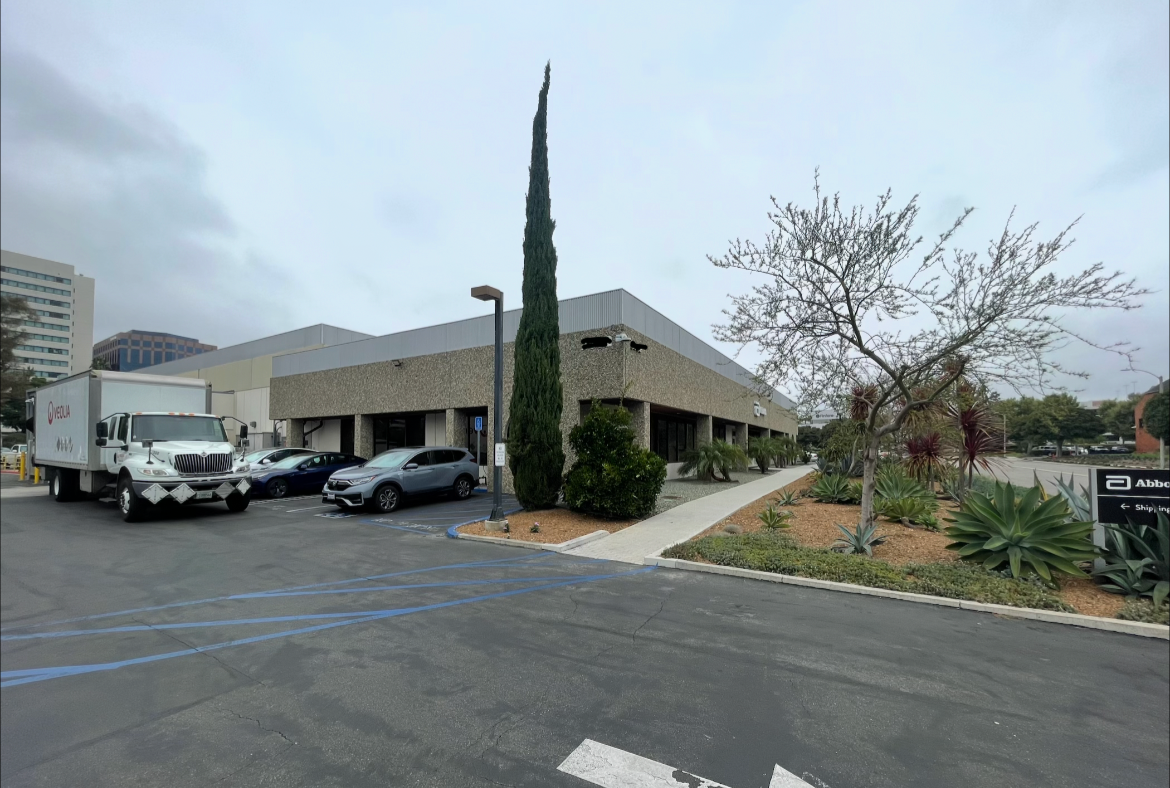Parking lot in front of a commercial building with a delivery truck, parked cars, and landscaped area with trees and succulents.