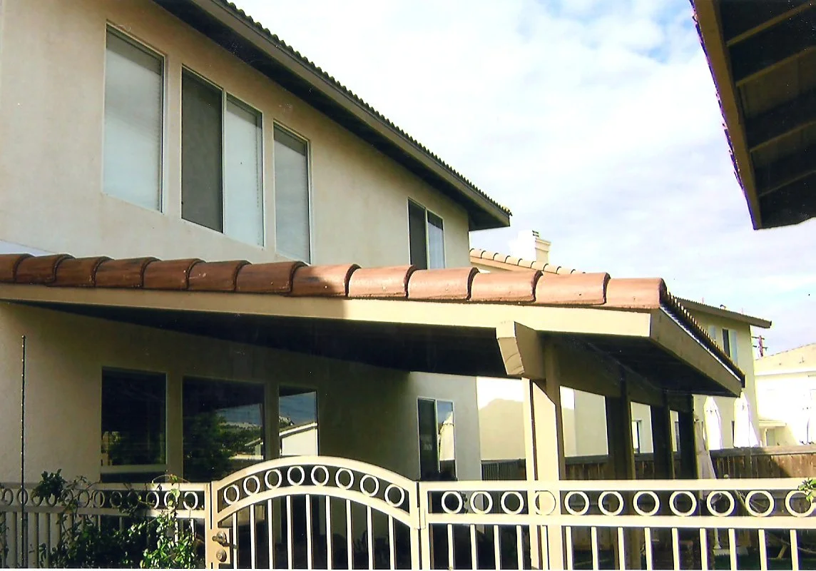 Two-story house with beige stucco exterior, large windows, and a red tile roof. A covered patio area has a gate with circular designs and some greenery.