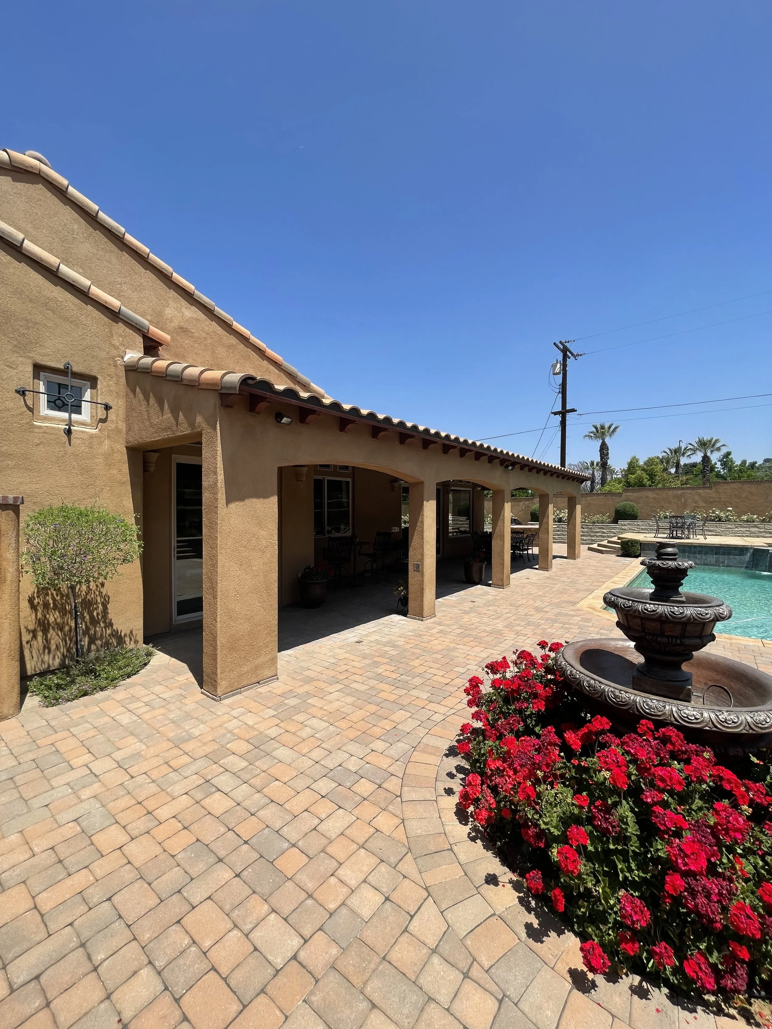Residential backyard with a swimming pool, a shaded patio area with chairs, potted plants, a water fountain surrounded by red flowers, and a stucco house wall with a small window and a windmill ornament.