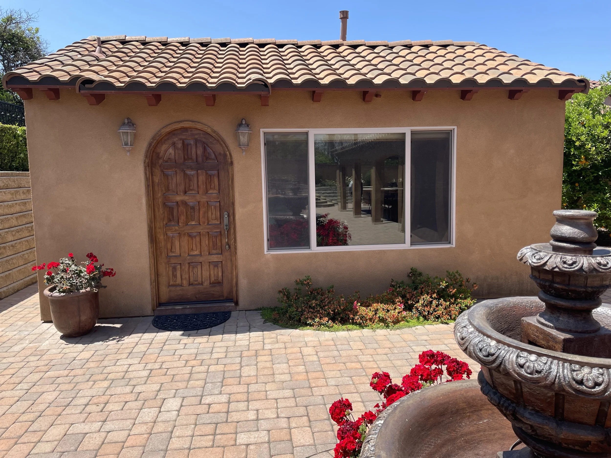 Small house with a tiled roof, beige stucco walls, a wooden arched front door, and a large window. There are decorative outdoor lanterns on each side of the door. The front yard has potted flowers, some bushes, and a decorative stone fountain with re