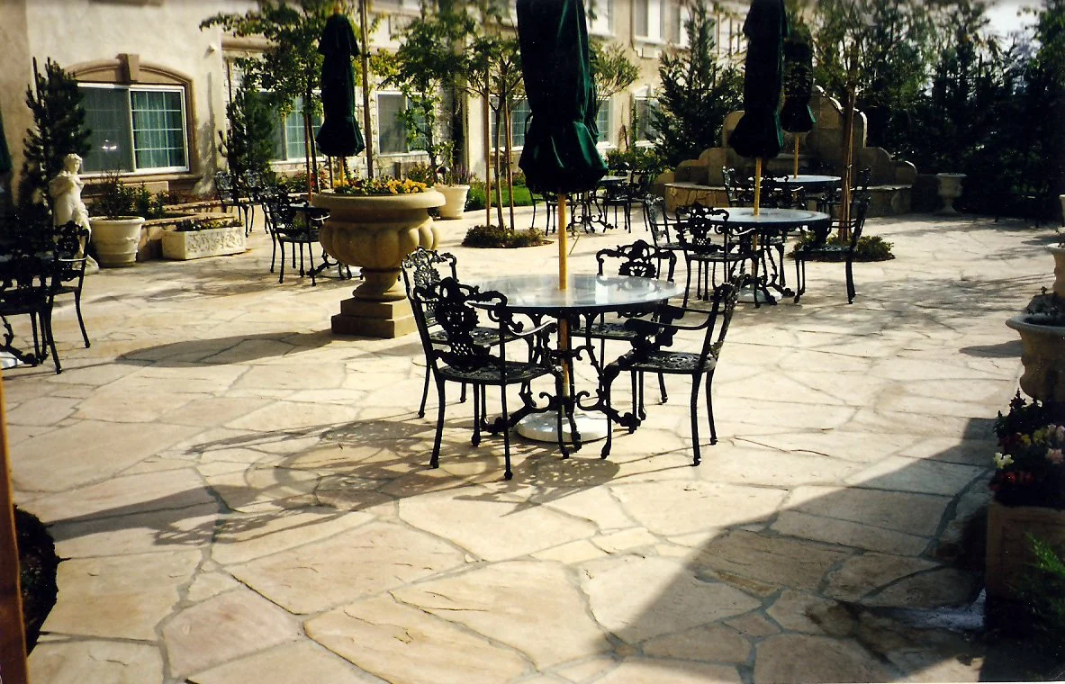 An outdoor patio with stone paving, black wrought iron tables and chairs, closed umbrellas, and decorative planters with flowers and plants.