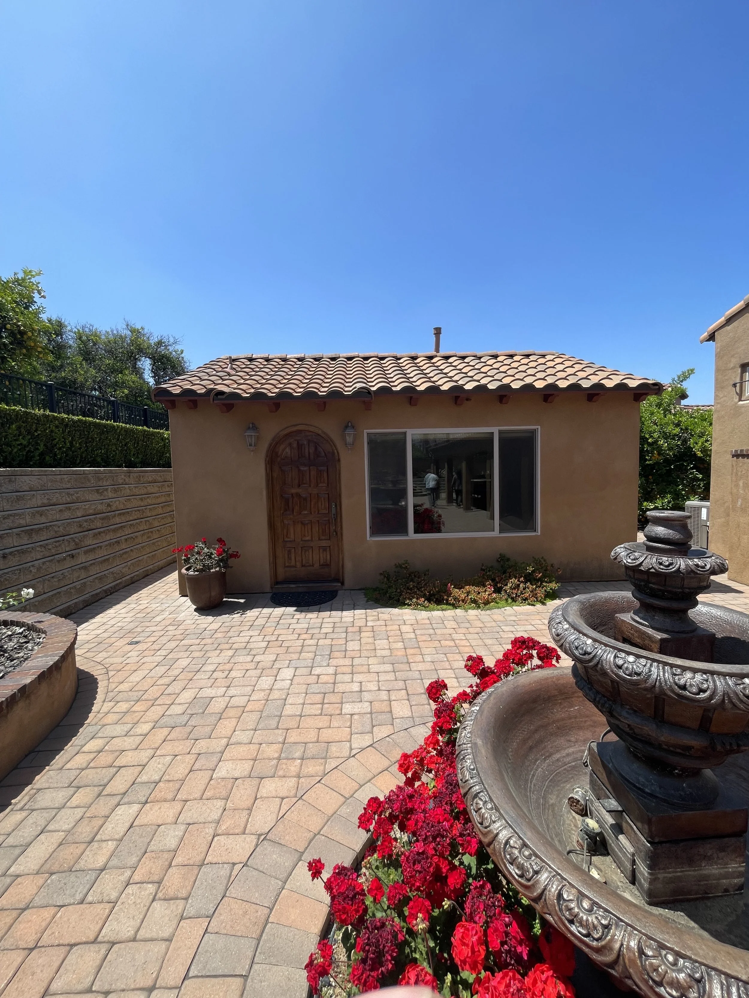 Small beige house with a red tile roof, a wooden door, and a large window. In the foreground, there is a decorative fountain surrounded by red flowers, and a paved patio area with a potted plant with red flowers. The sky is clear and blue.