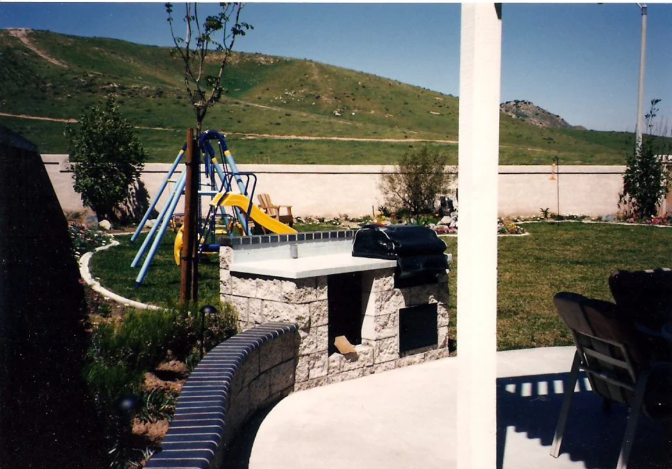 A backyard with a small colorful playground slide, a stone barbecue grill, and a white wall in the background. There are some trees, a grassy lawn, and a mountain in the distance in Corona, CA.