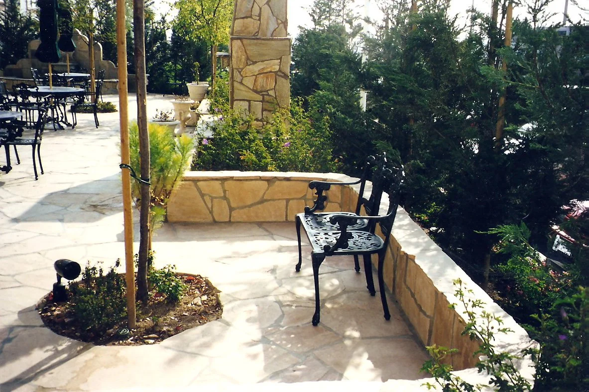 Outdoor patio area with stone flooring, black metal chairs, a stone planter with plants, a young tree in a circular bed, and greenery in the background.