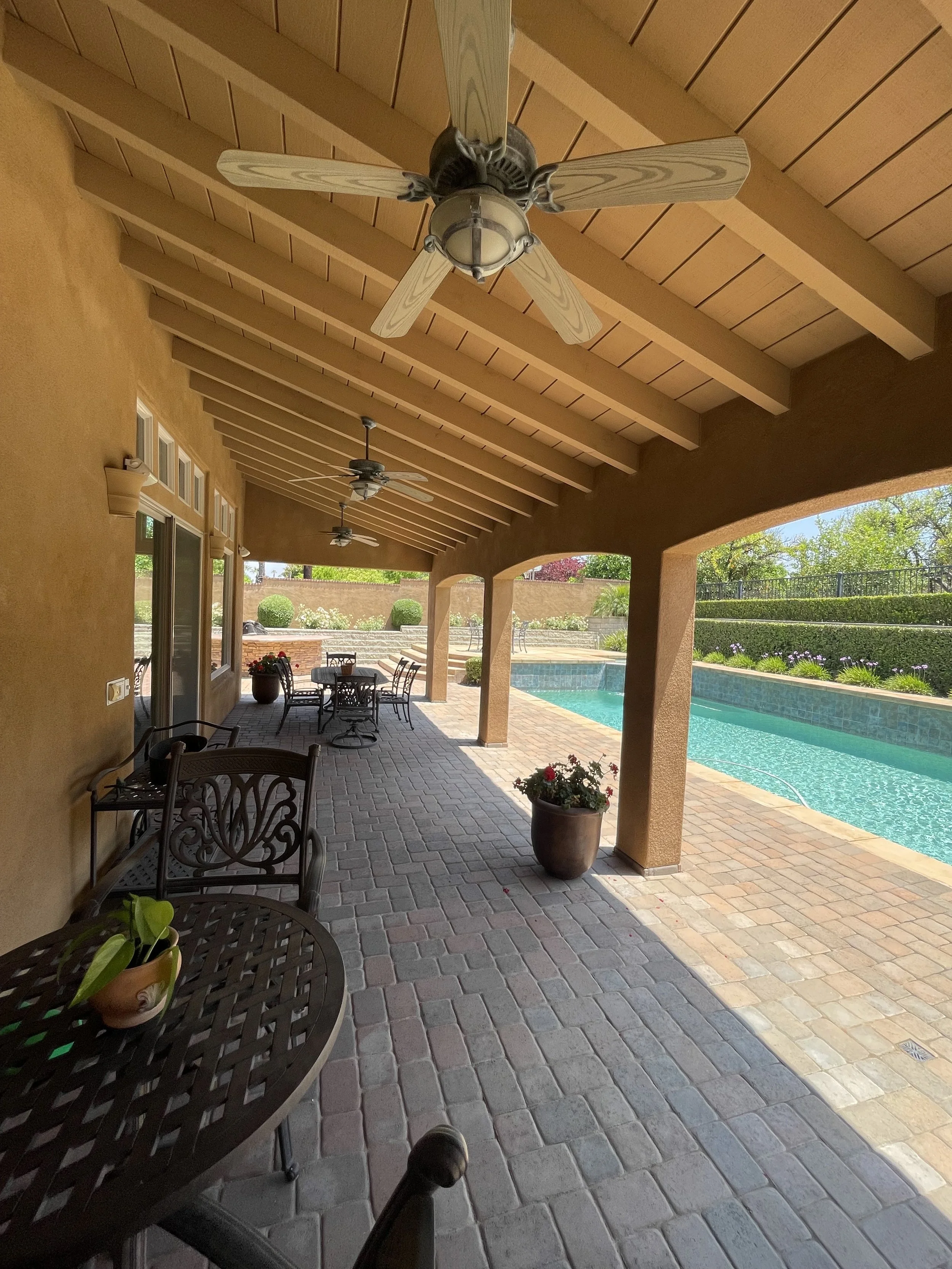 Covered patio area with brick flooring, ceiling fans, potted plants, and outdoor furniture overlooking a swimming pool and garden.