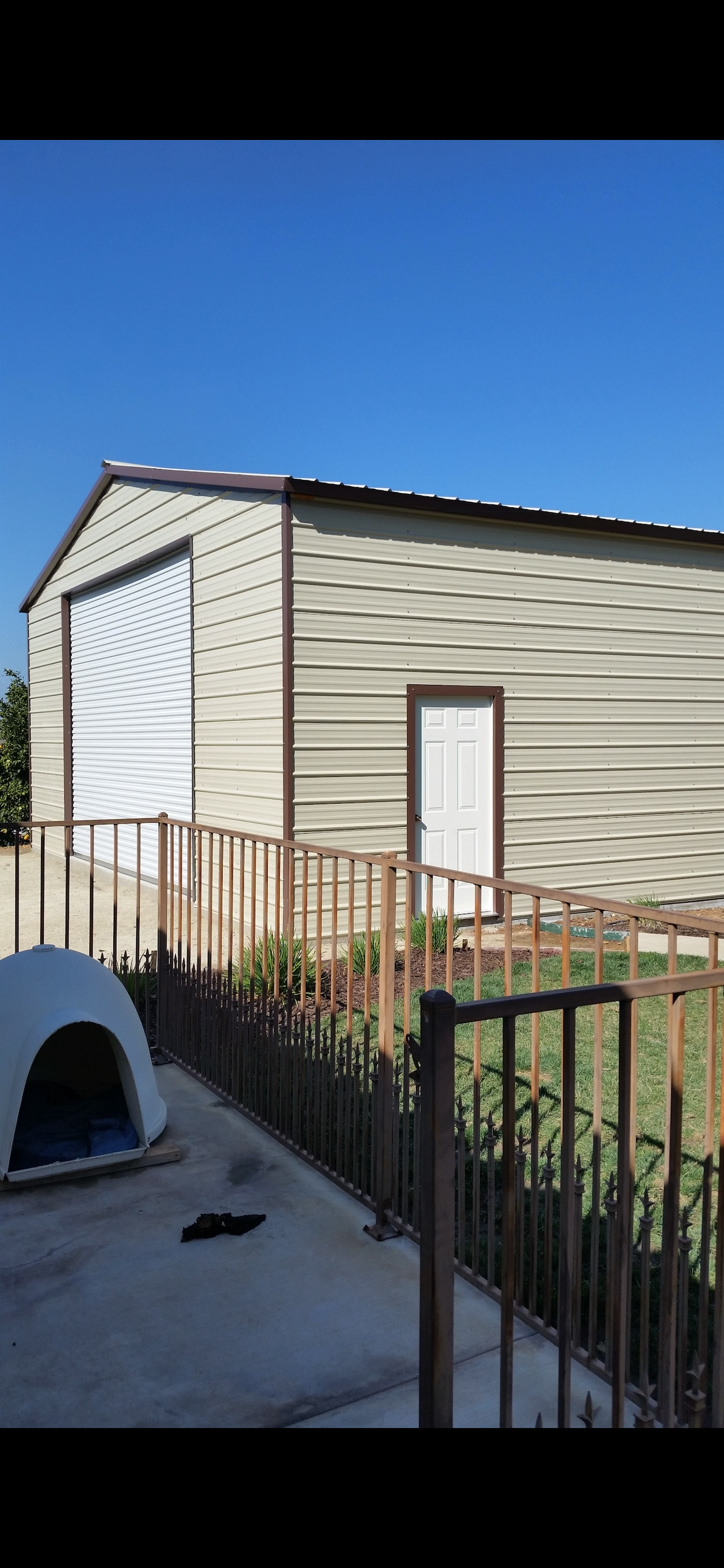 Photo of a beige metal storage building with a white roll-up door and a white side door, surrounded by a metal fence, with a small green grass area and plants in front, and a doghouse in the foreground.