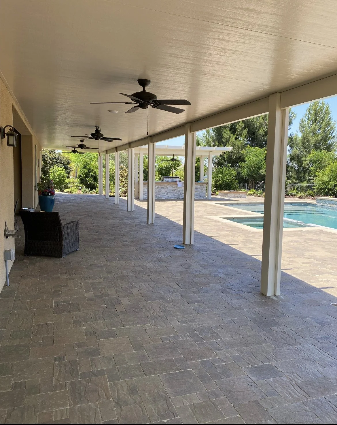 Covered patio with ceiling fans, a wicker chair, and a large blue planter, overlooking a backyard with a swimming pool, lush green trees, and a stone outdoor kitchen area.
