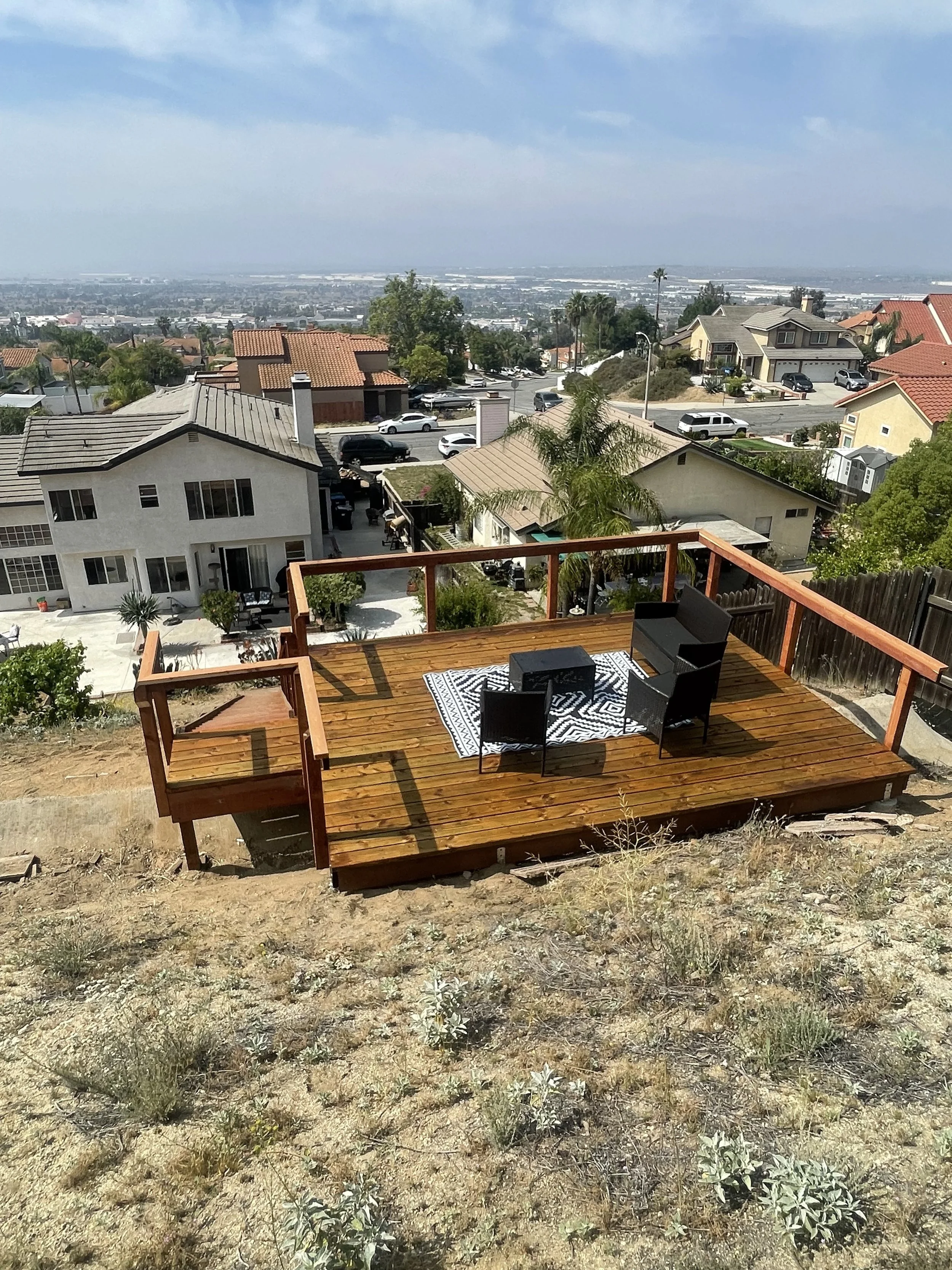 Wooden deck with three black chairs, a small black table, and a white and black patterned rug, overlooking a suburban neighborhood with houses and trees in Moreno Valley