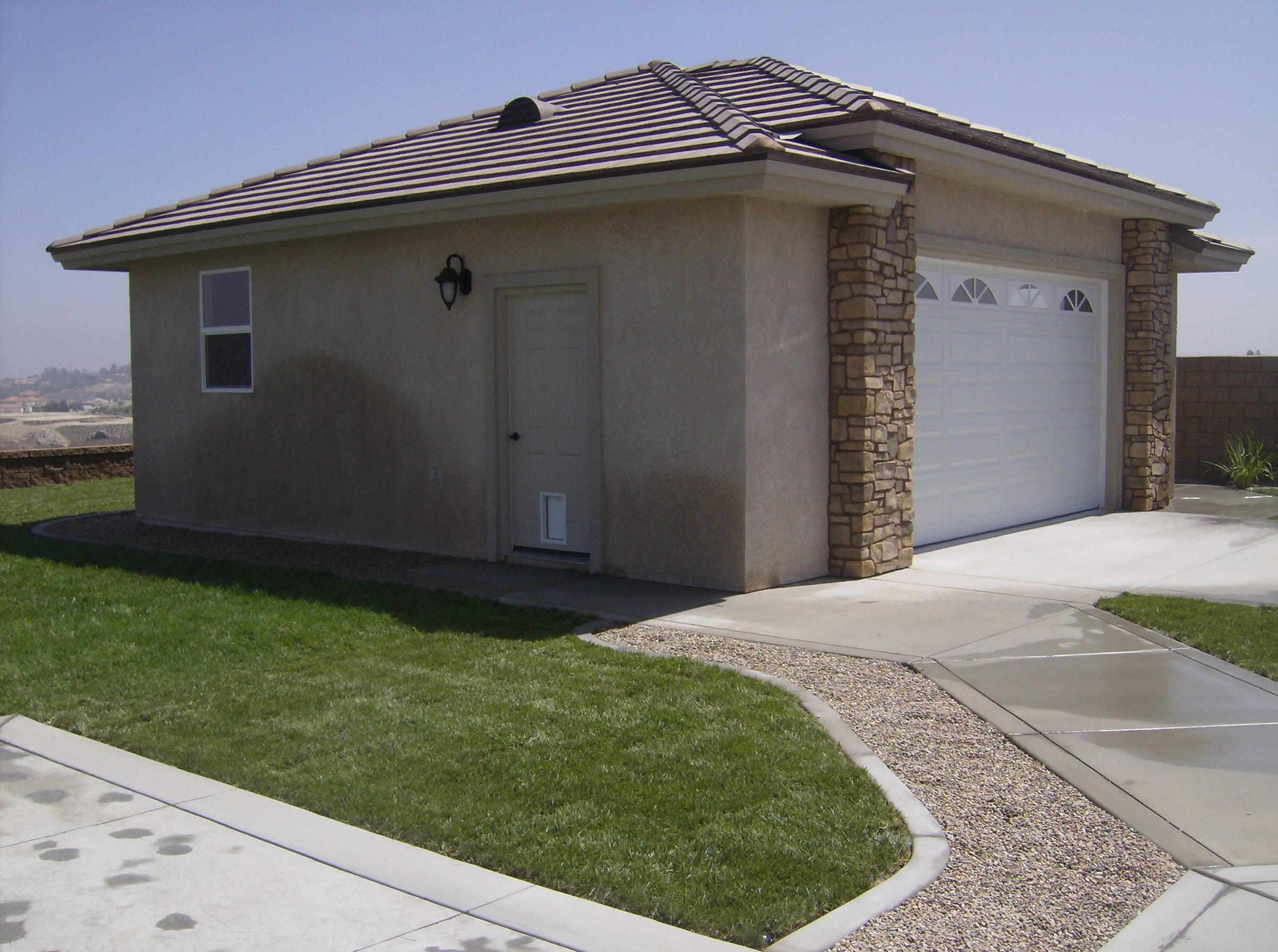 A small beige house with a tiled roof, a white garage door, and a side door with a black wall-mounted lantern, surrounded by green lawn and concrete driveway.