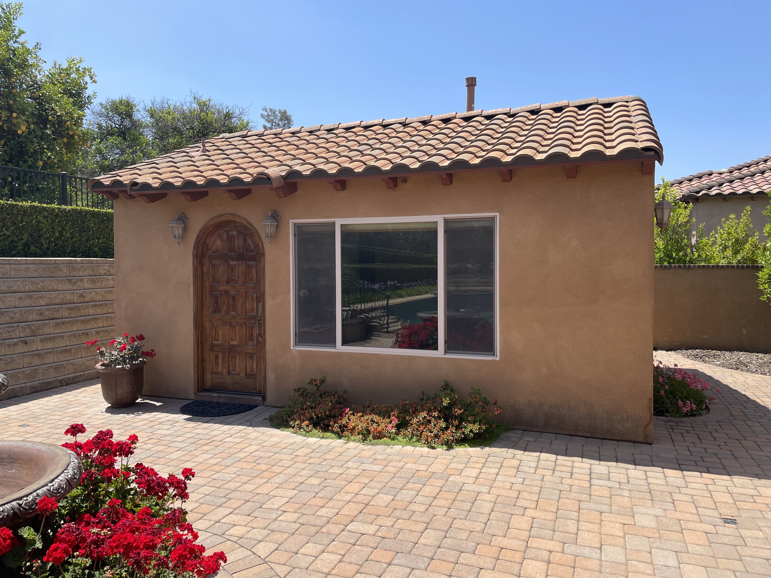 Small tan stucco house with a brown tiled roof, a wooden arched front door, a large window, outdoor wall-mounted lanterns, and red flowers in pots and garden beds in a paved courtyard.
