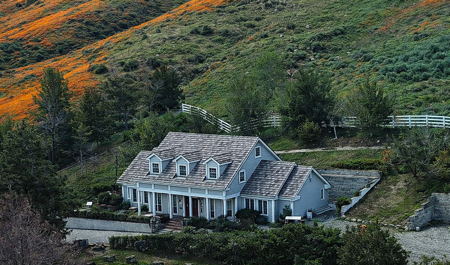 A house in Gavilan Hills, Riverside with gray siding and a shingled roof situated on a hillside with trees and green vegetation, and a mountain in the background.