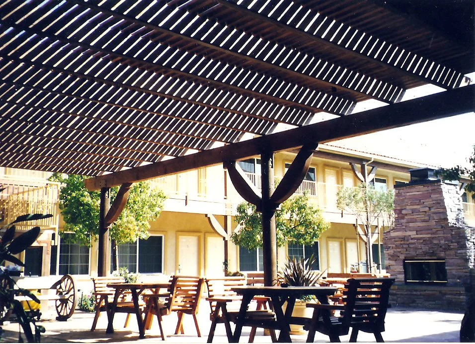 Outdoor patio area with wooden tables and chairs, a stone fireplace, trees, and a building in the background, shaded by a slatted wooden pergola.