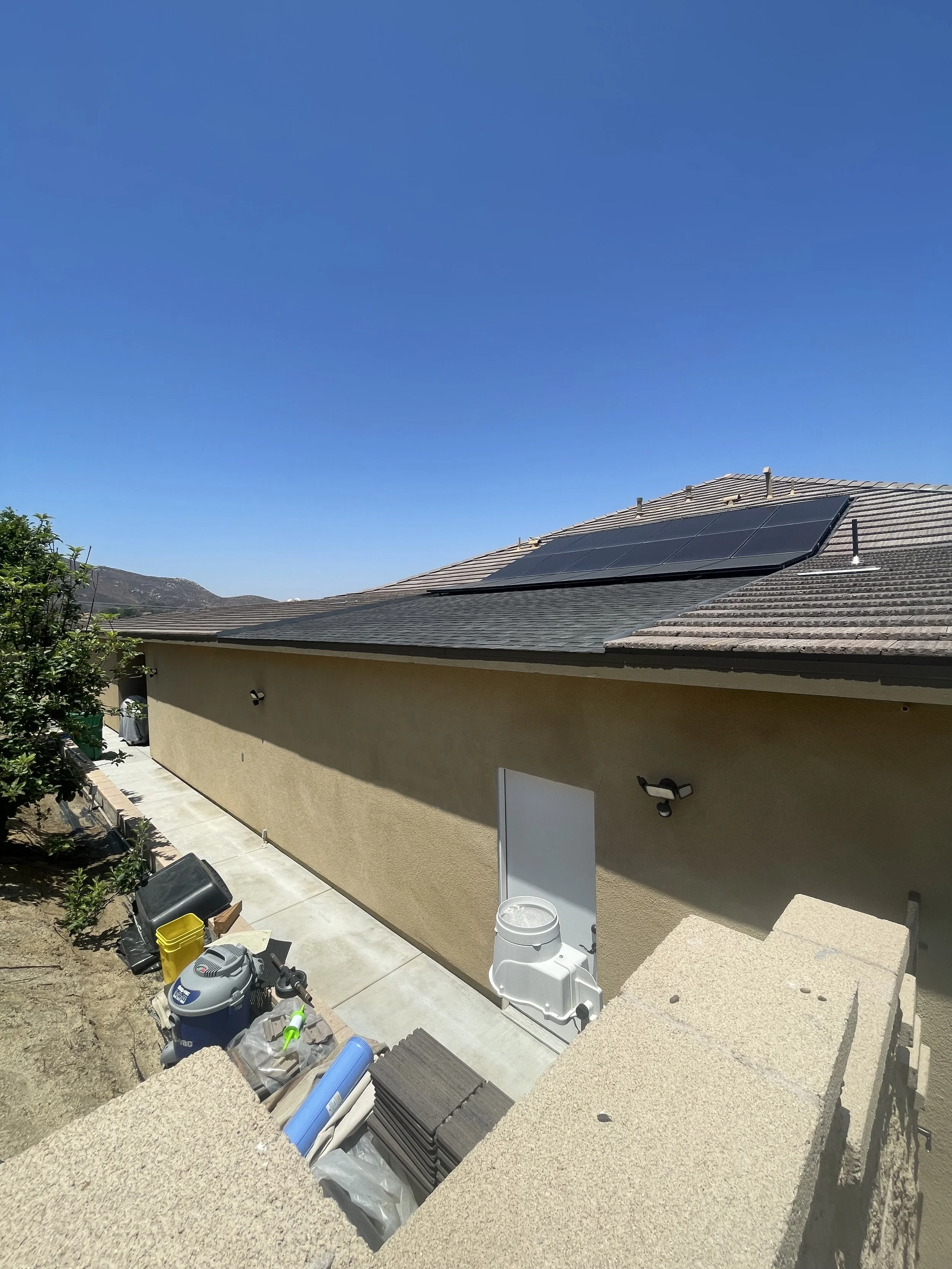 View of a house with solar panels on the roof, beige exterior wall, outdoor light fixtures, a white door with a dog bowl, and construction supplies on a concrete walkway. Clear blue sky.