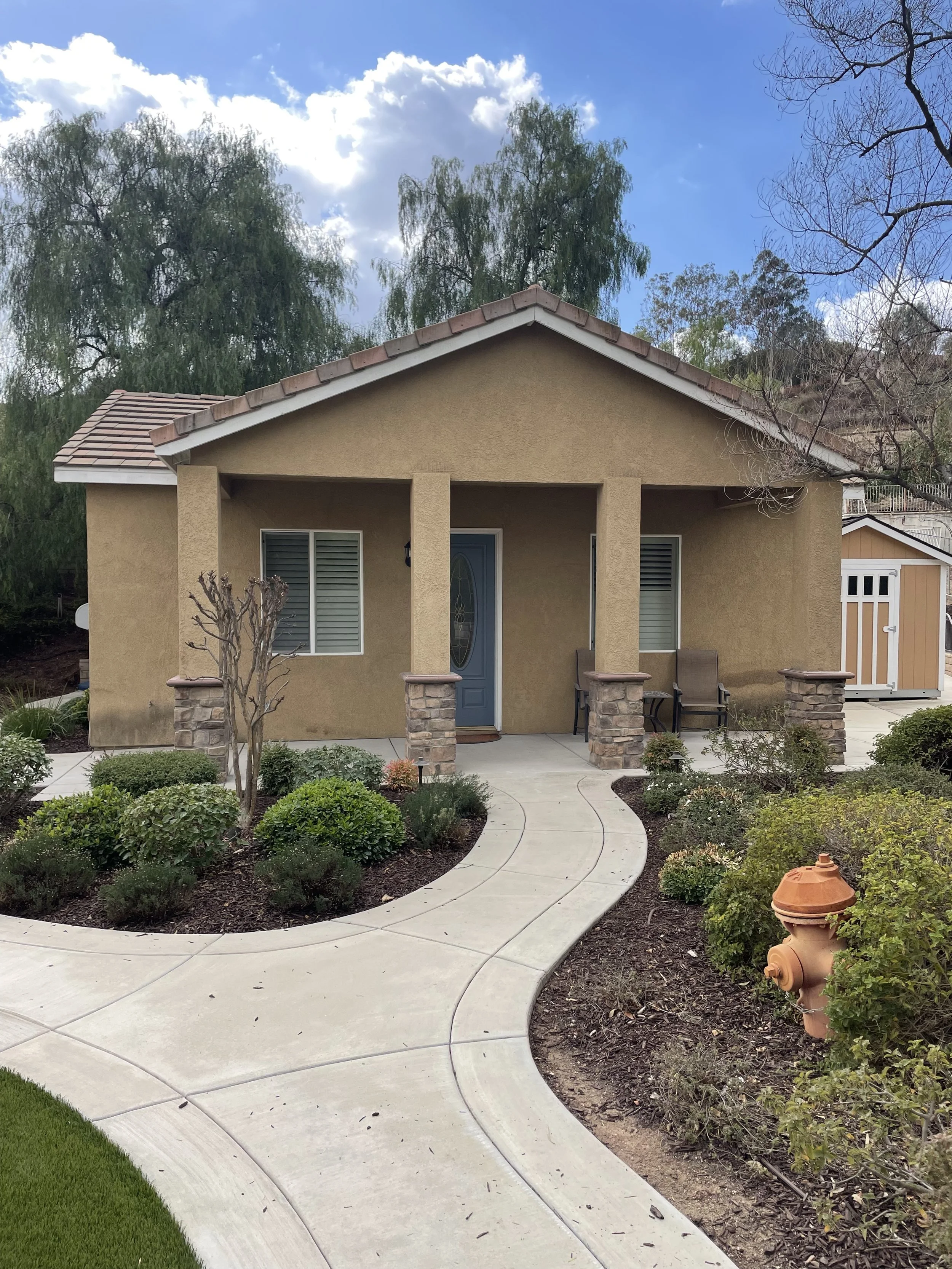 Front view of a small beige house with a front porch and a curved sidewalk leading to the door, surrounded by landscaped bushes and trees under a partly cloudy sky.