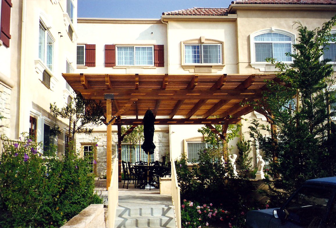 A residential building with a wooden pergola outdoor patio, surrounded by greenery and flowers, with a table and chairs underneath the pergola.