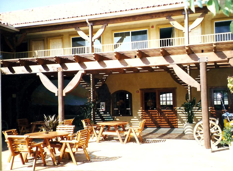 Outdoor patio area with wooden tables and chairs, potted plants, and a wooden balcony with doors and windows, part of a building with a yellow exterior.