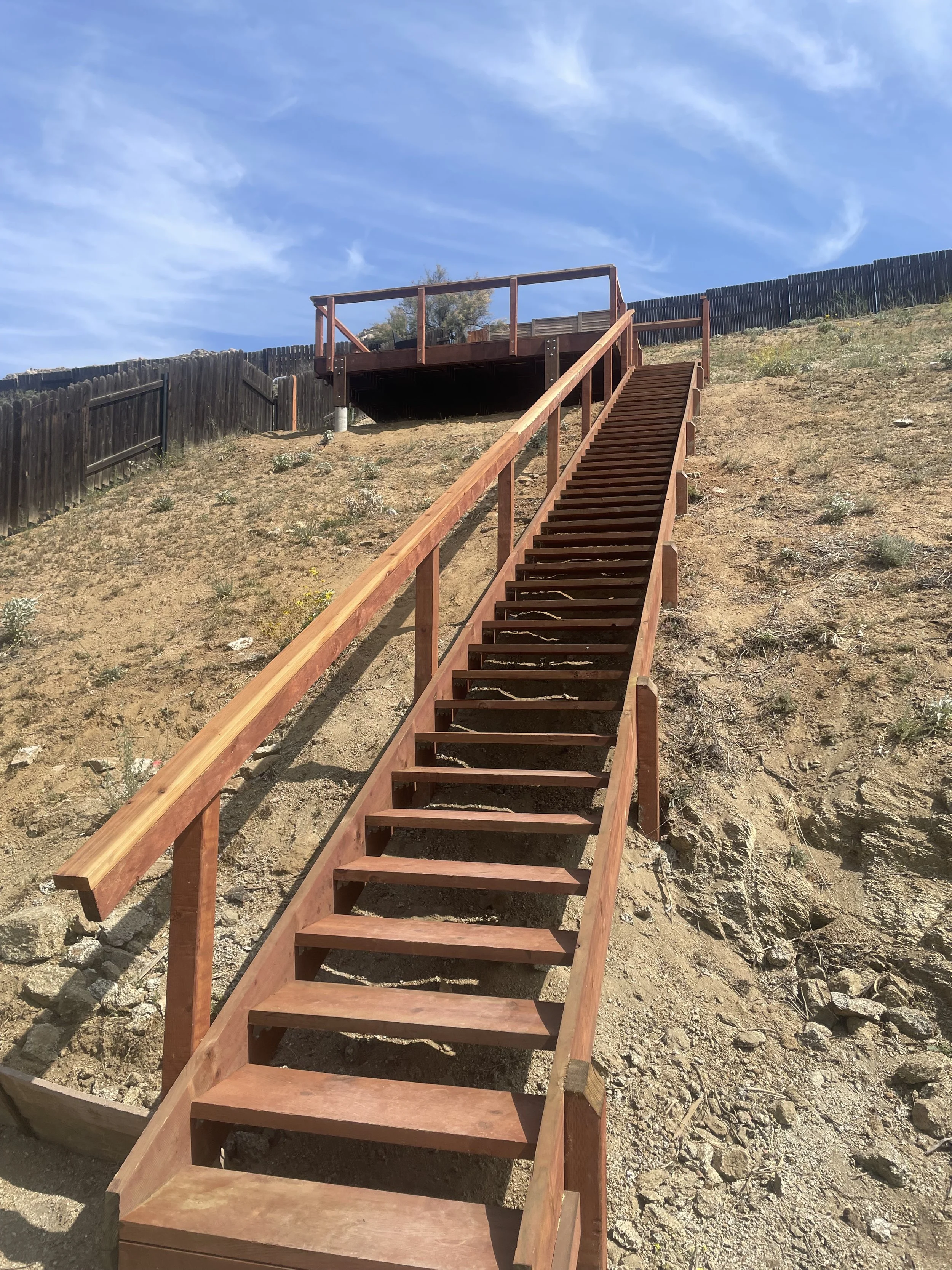 Wooden staircase leading up a dirt slope to a deck with a glass railing, set against a bright blue sky in Moreno Valley.