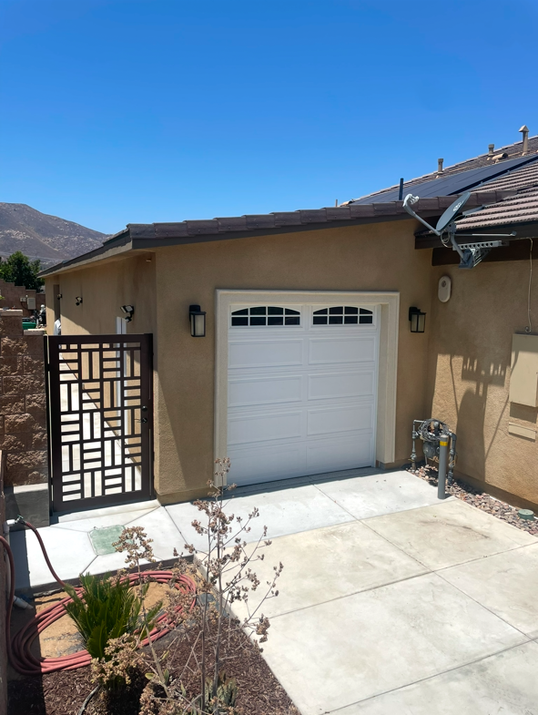 A beige house with a white garage door, outdoor wall lights, and a wooden gate. A satellite dish and utility meters are visible, with a garden hose and plants in the front yard. Blue sky and mountains in the background.