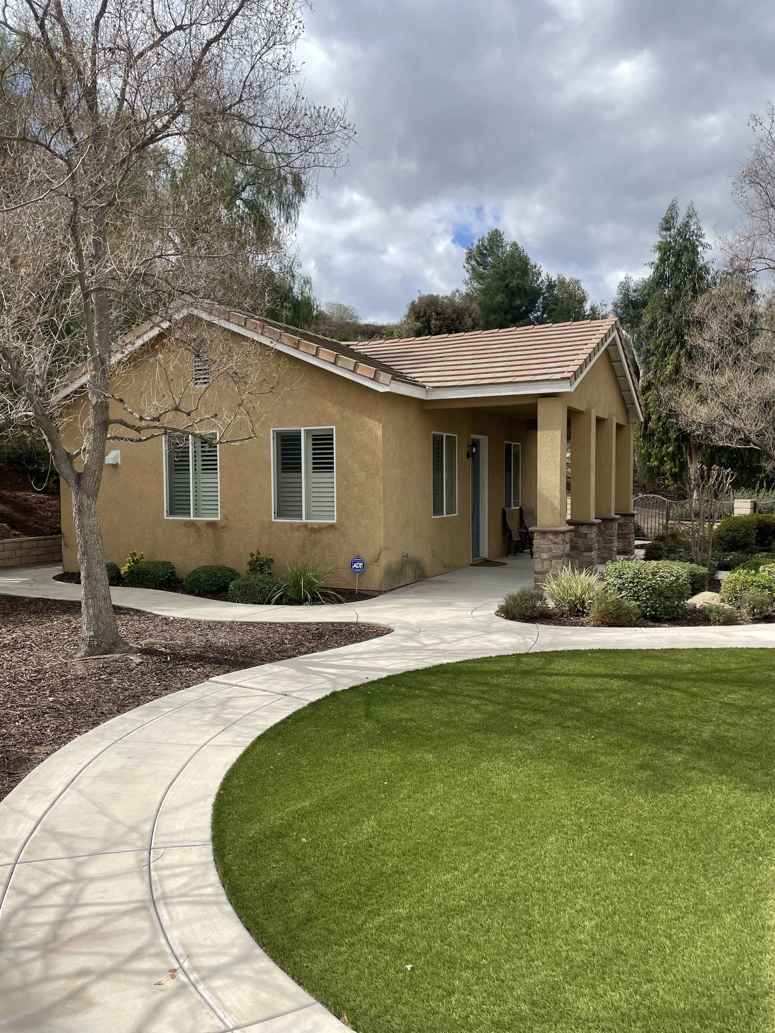 Single-story house with yellow exterior walls, a terracotta tiled roof, and a front porch with stone pillars. Well-maintained lawn, curved walkway, and landscaped garden with bushes and trees, some without leaves. Overcast sky.