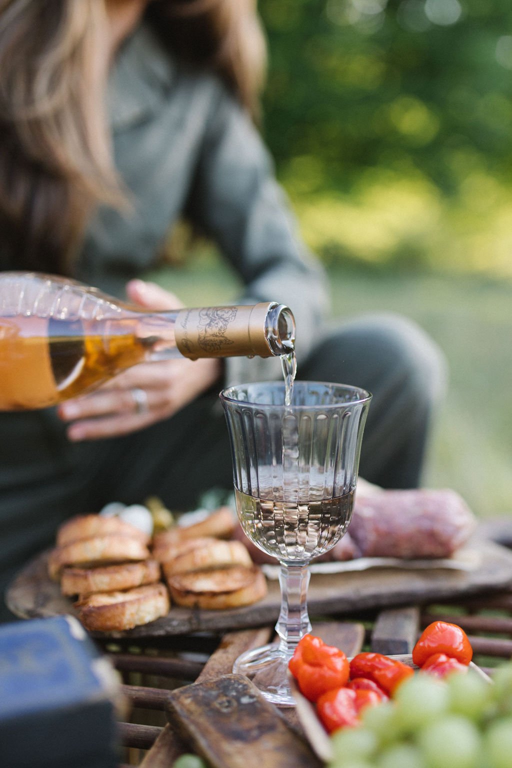 Person pouring rosé wine into a glass outdoors with snacks including grapes, tomatoes, and pastries on a wooden table.