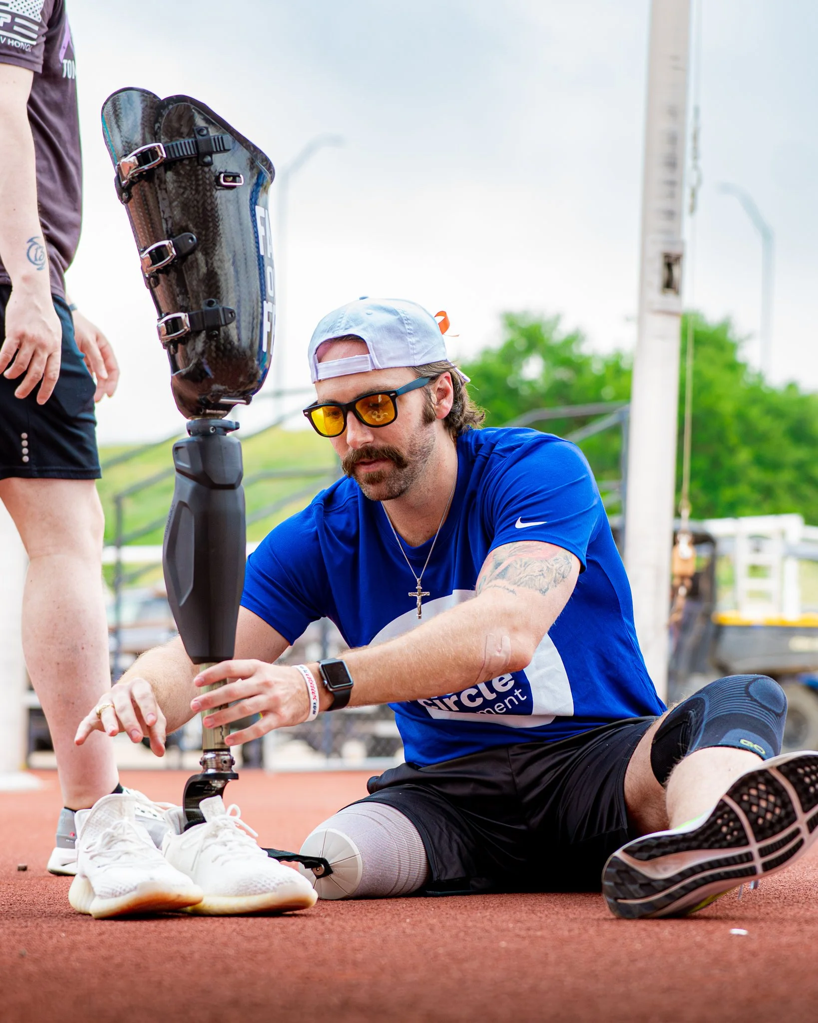 A man with a prosthetic leg, sunglasses, a white cap, and a mustache, wearing a blue t-shirt, sitting on the ground on a sports track, attaching his shoes to a prosthetic running blade.