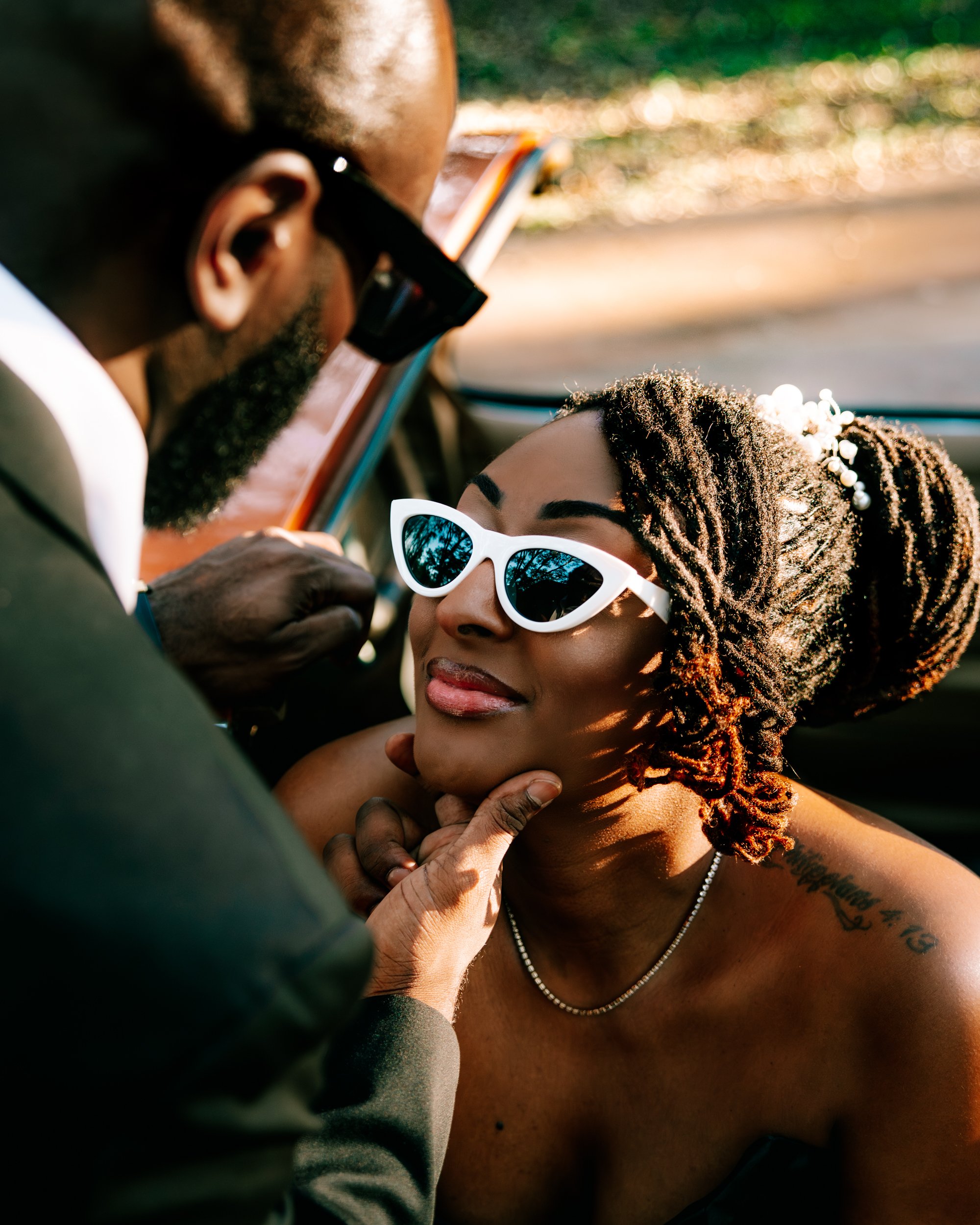 A man and woman sitting in a car, with the man adjusting the woman's white sunglasses. The woman has dreadlocks adorned with pearls, a tattoo on her shoulder, a pearl necklace, and is smiling softly. Sunlight is shining through the car window.