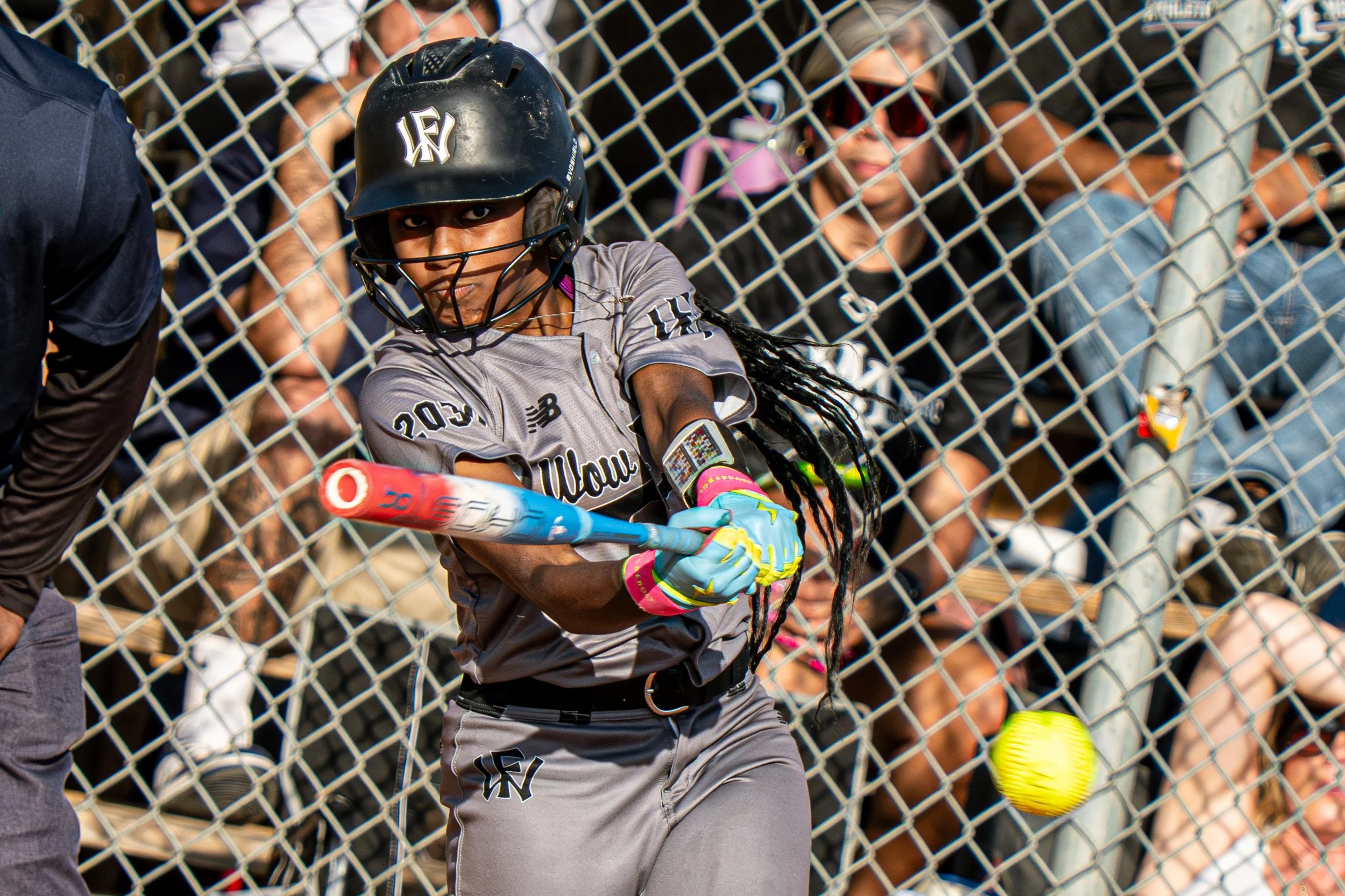A female softball player in a gray uniform and helmet is batting, with a yellow-green softball approaching her bat. She is wearing brightly colored gloves and is in front of a chain-link fence with spectators watching in the background.