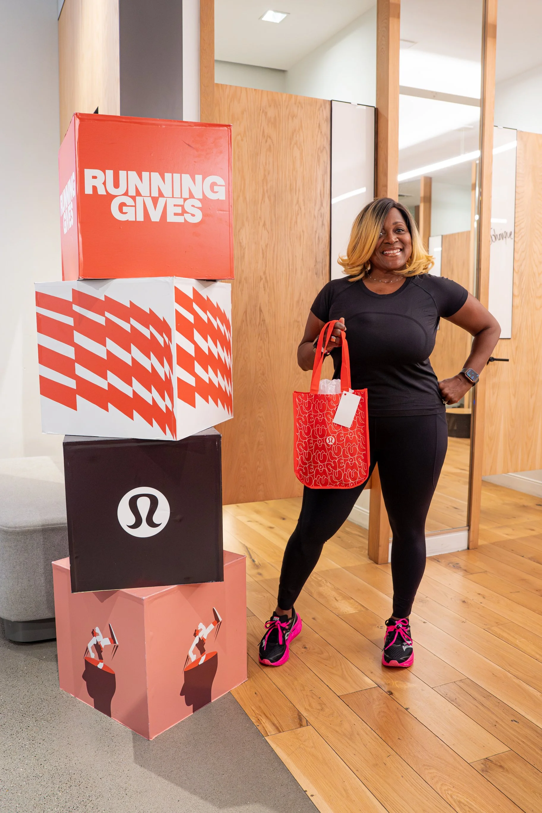 A woman wearing a black athletic outfit and pink running shoes smiling while holding a red tote bag, standing next to colorful boxes with fitness-related logos and slogans.