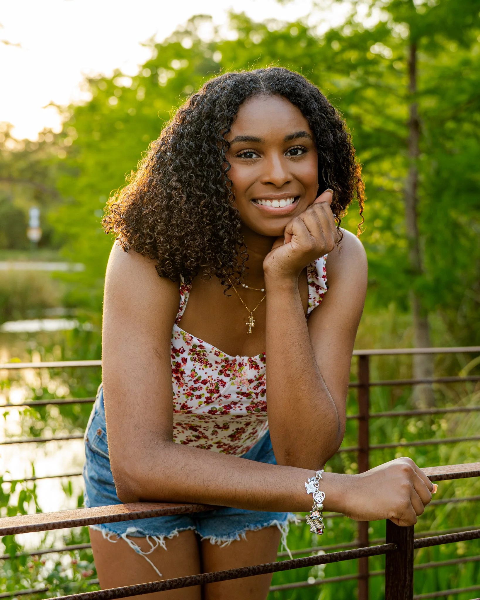 A young woman with curly hair smiling, resting her chin on her hand, outdoors near a body of water with green trees in the background.