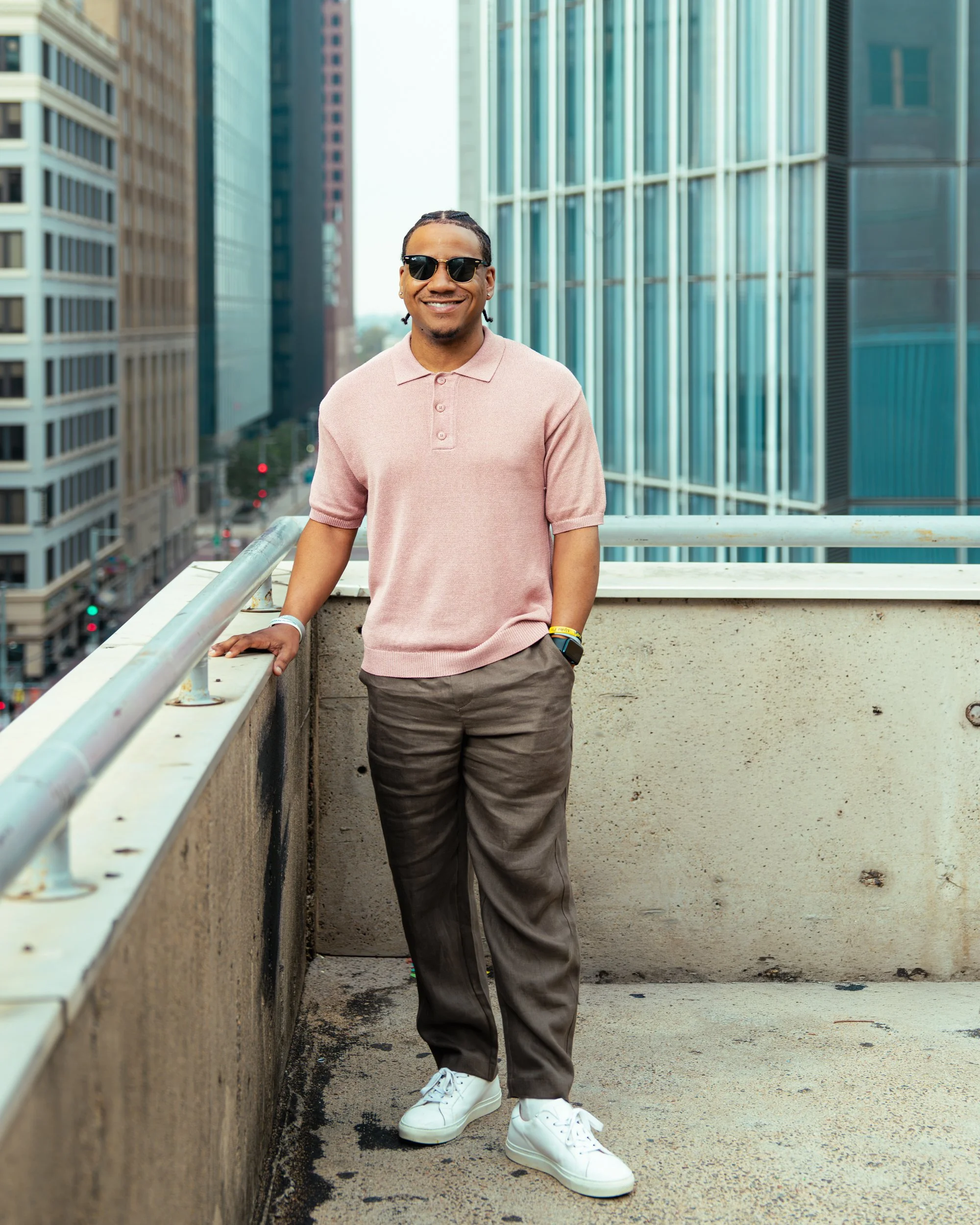Man with sunglasses standing on a rooftop balcony in an urban area, wearing a pink polo shirt, gray pants, and white sneakers, smiling, with tall buildings in the background.