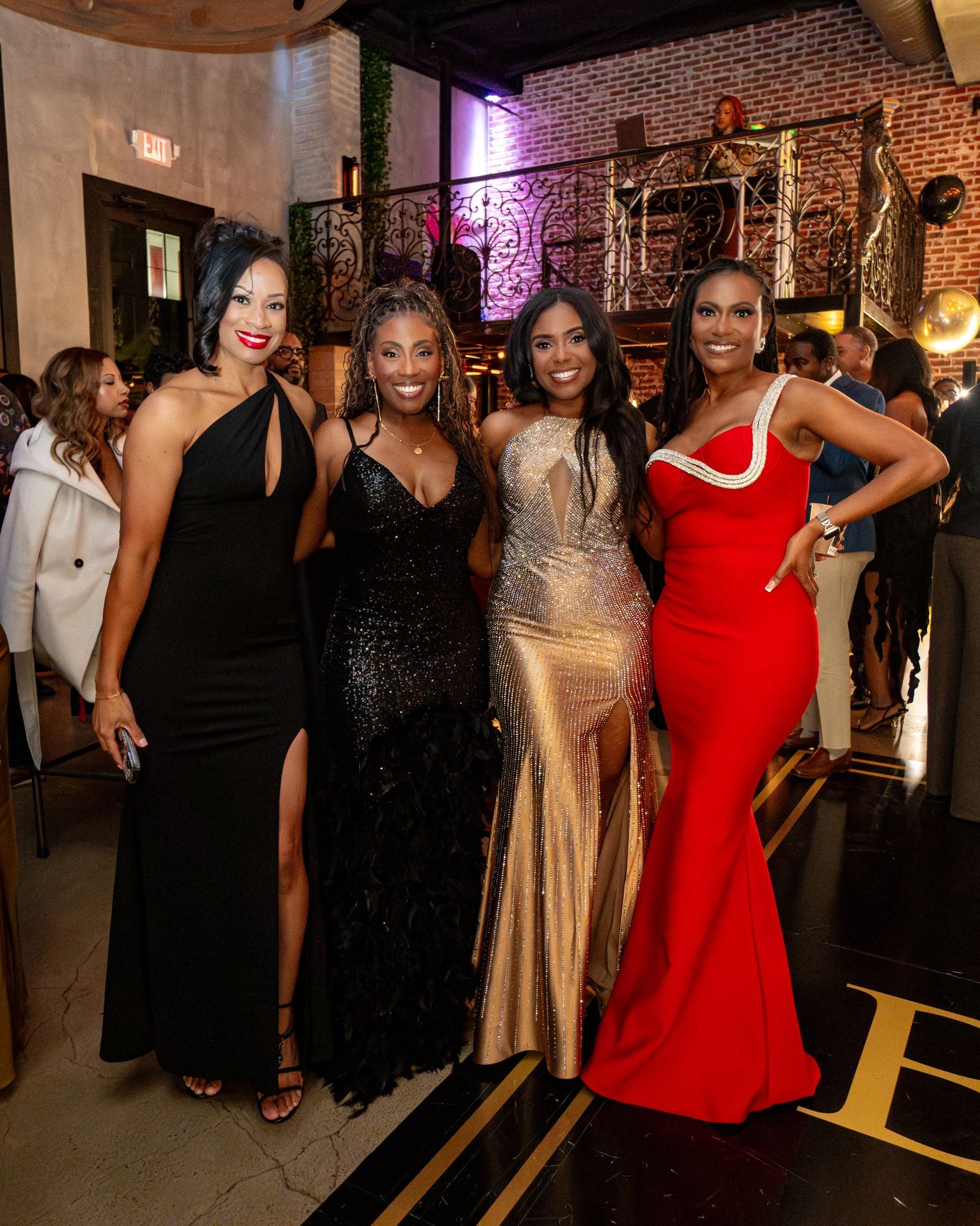 Four women dressed in elegant evening gowns posing together at a formal event in a decorated venue with a brick wall and a DJ booth in the background.