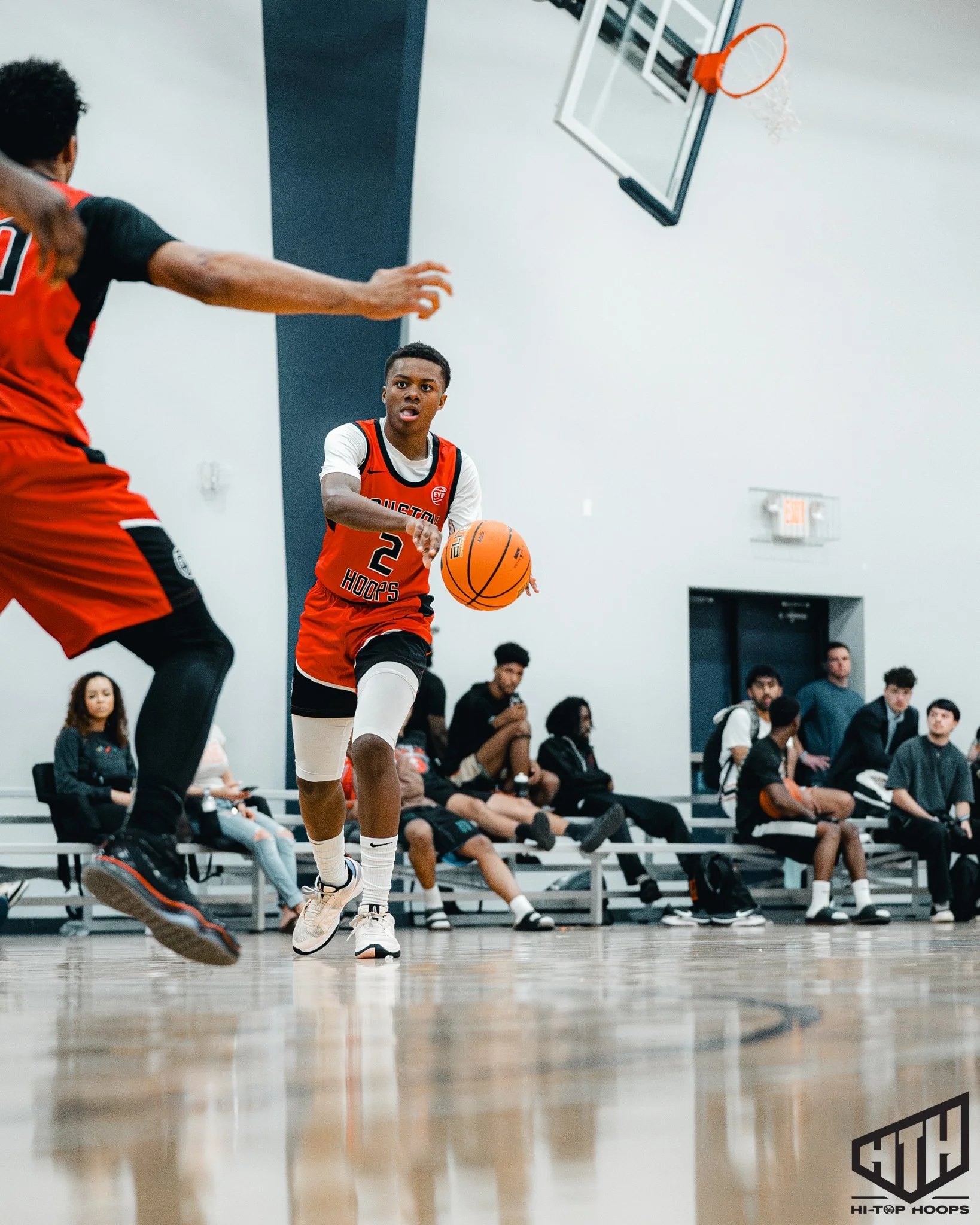 Young basketball player in a red and black jersey dribbling the ball on the court during a game, with spectators sitting on benches in the background.