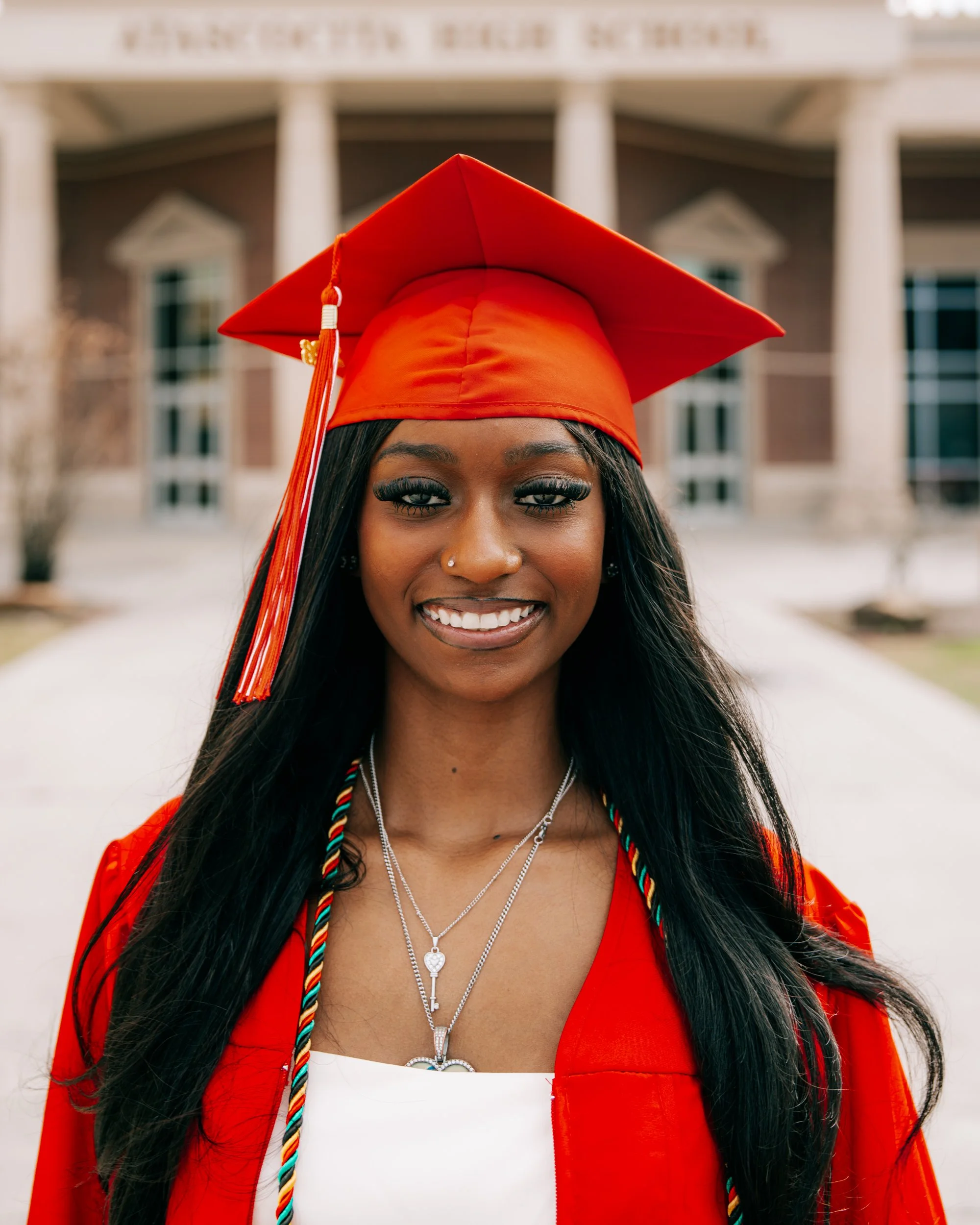 Smiling young woman in red graduation gown and mortarboard posing outdoors in front of a school building.