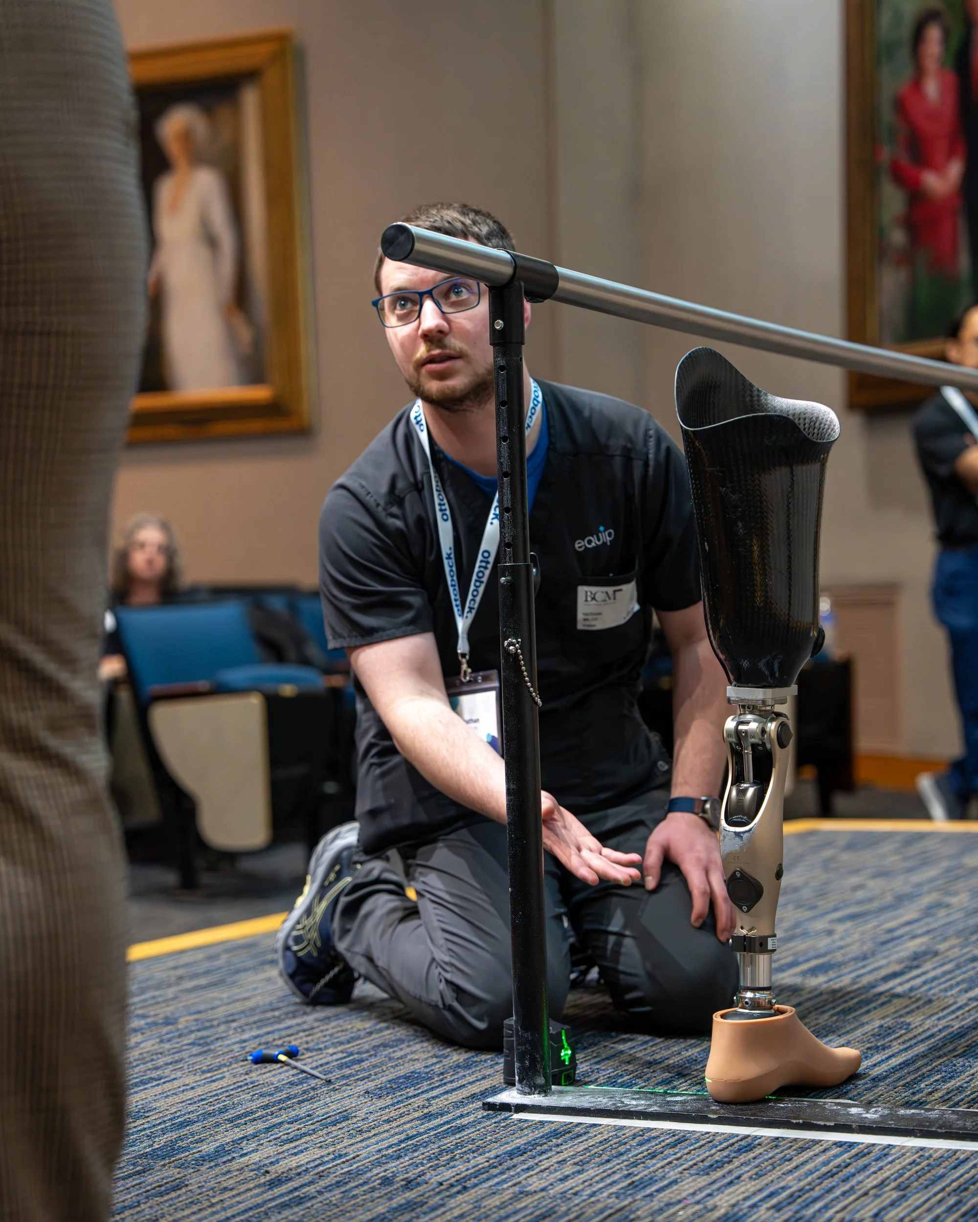 Man kneeling on the floor observing a robotic prosthetic leg during a demonstration or competition, with paintings hanging on the wall behind him.