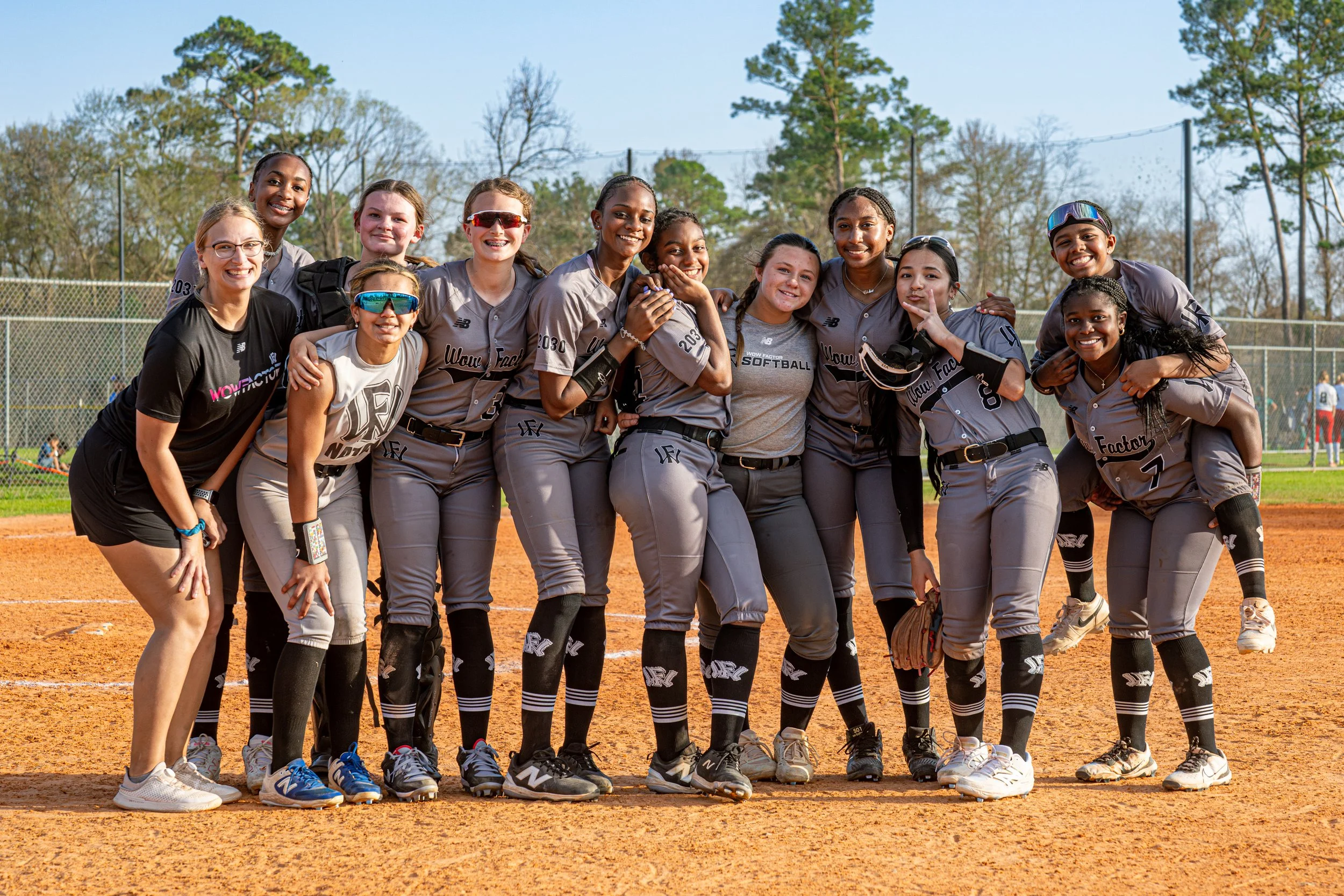 A group of twelve young women, part of a softball team, standing together on a baseball field and posing for a team photo on a sunny day.