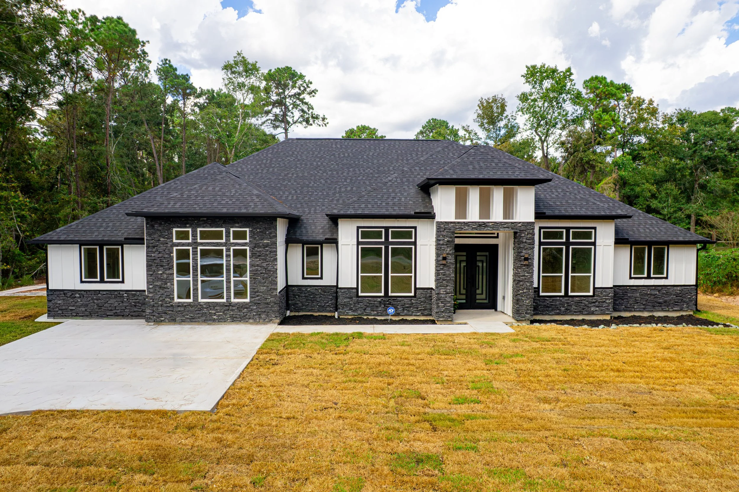 Modern house with black tiled roof, white paneling, and black stone facade, surrounded by trees, with a concrete driveway and grass lawn.