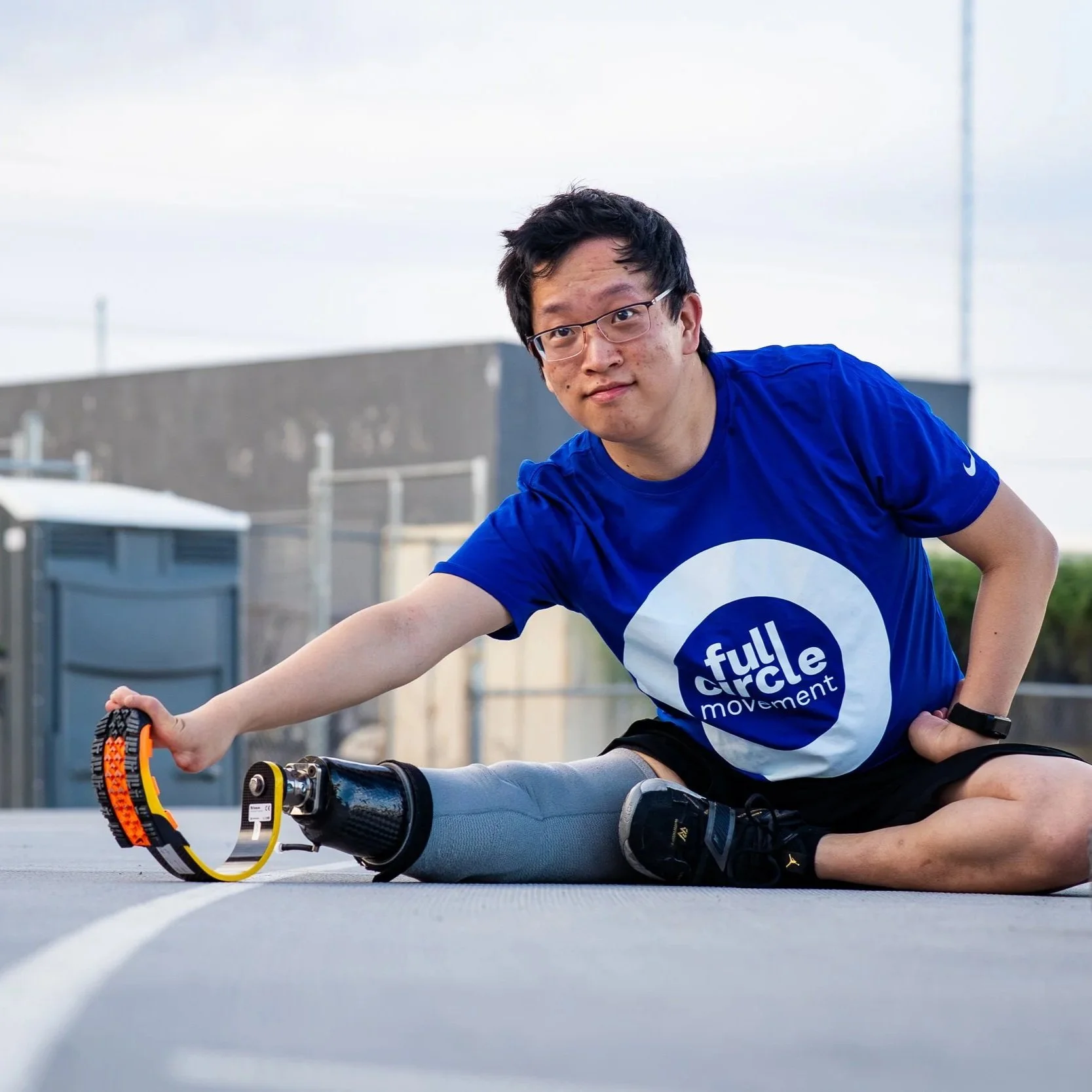 Young man with glasses, wearing a blue t-shirt with 'full circle movement' logo, stretching on a rooftop with a prosthetic leg, holding a massage roller.