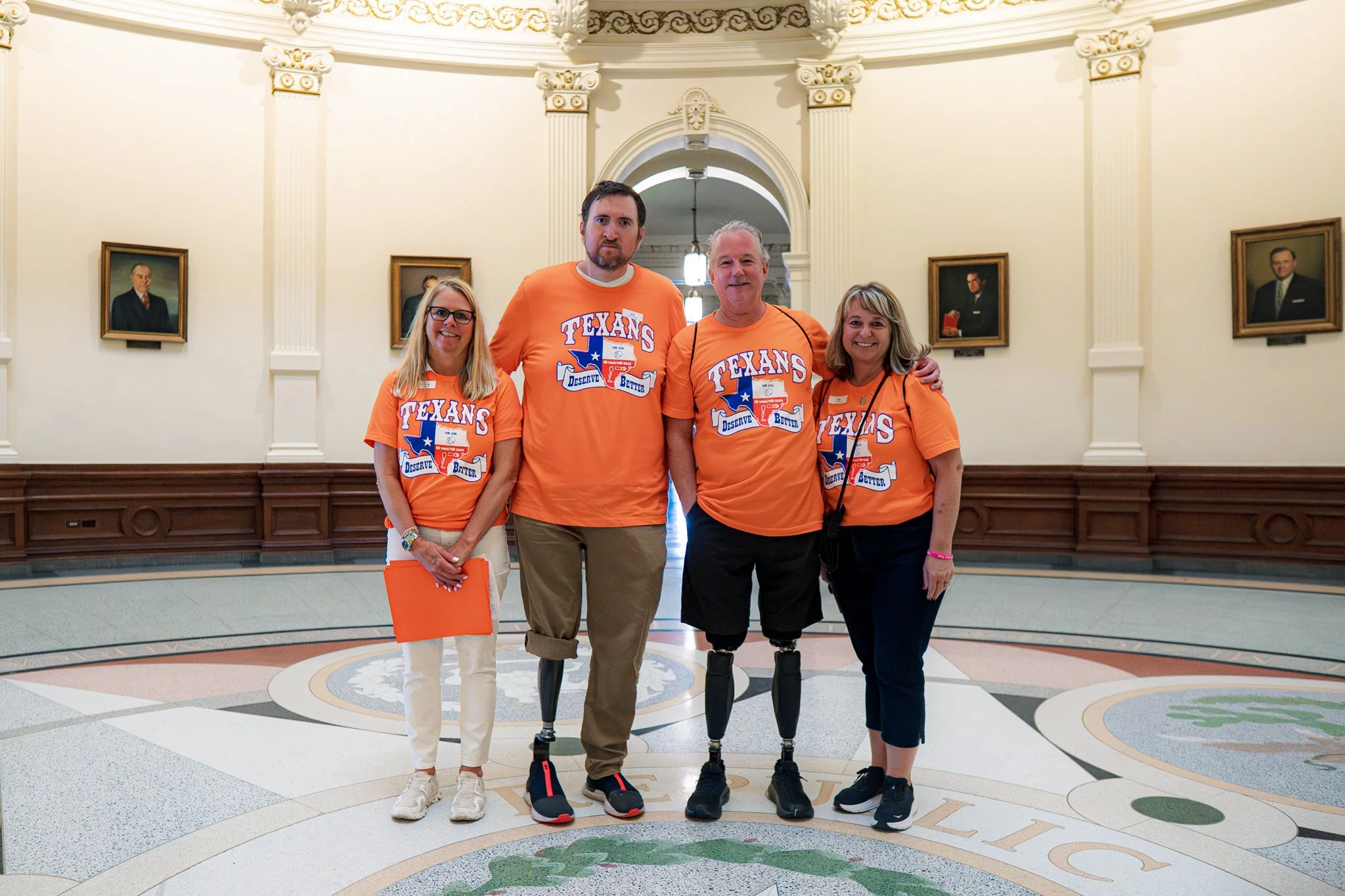 Four people standing together in a grand, historic building with portrait paintings on the wall. All are wearing orange T-shirts with a Texas-themed graphic and text. The two women and one man have prosthetic or robotic legs. They are smiling and leaning slightly towards each other.