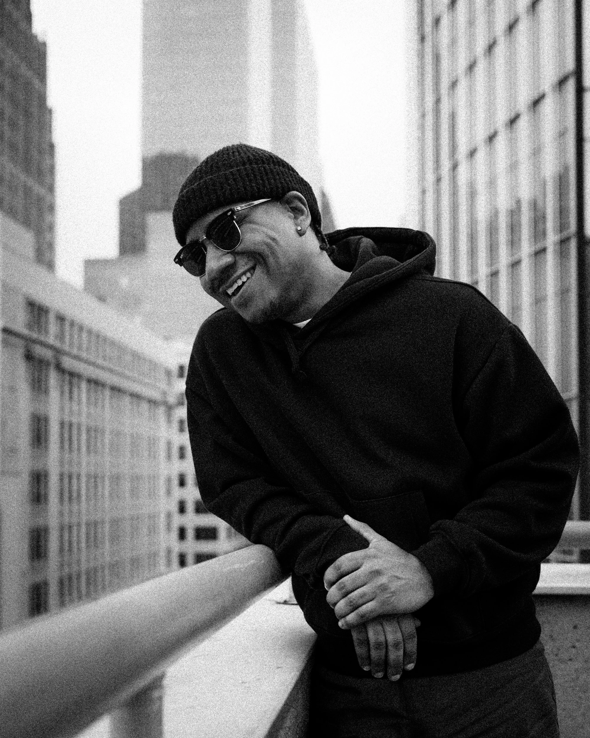 A man in a beanie hat and sunglasses leaning on a balcony railing in an urban cityscape, smiling in black-and-white.