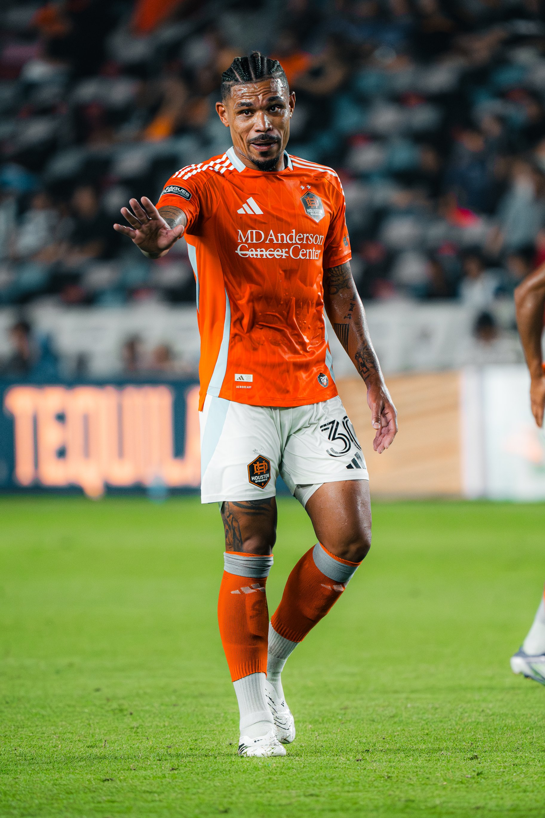 A soccer player in an orange Houston Dynamo jersey and white shorts on the field, waving with his right hand, during a game, with a stadium crowd in the background.