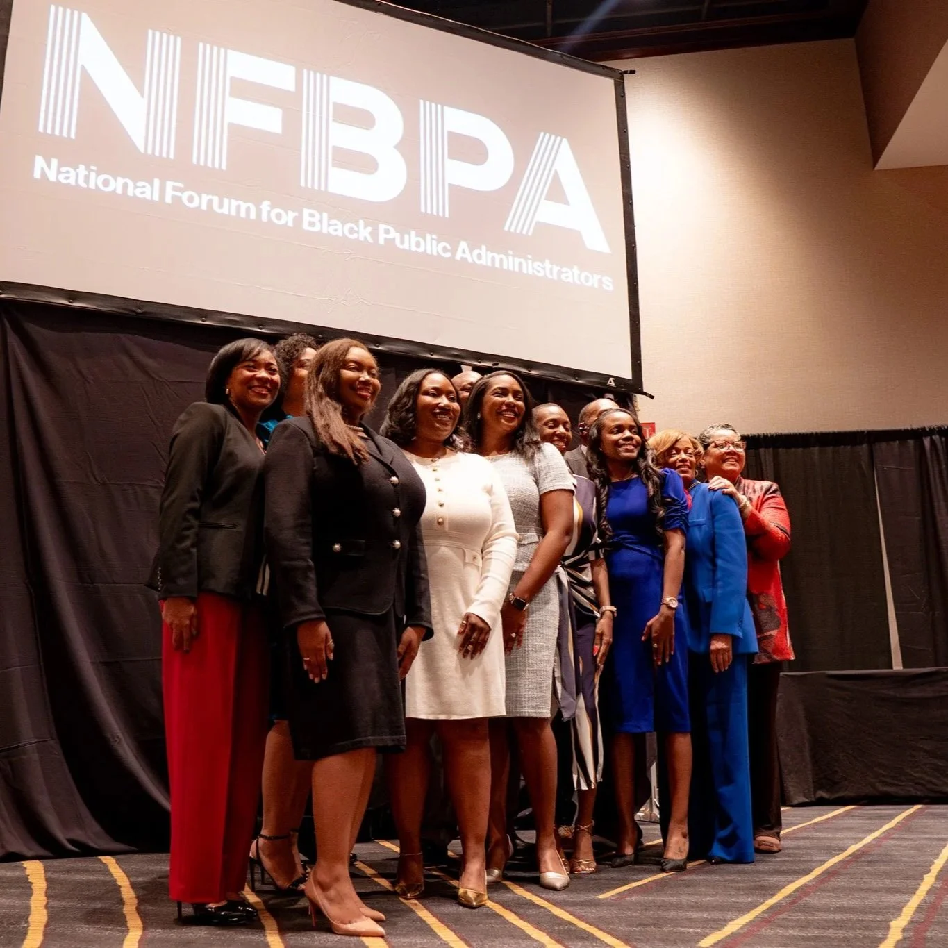 A group of women standing on stage at an event, smiling, in front of a large screen displaying the logo and name of the National Forum for Black Public Administrators (NFBPA).