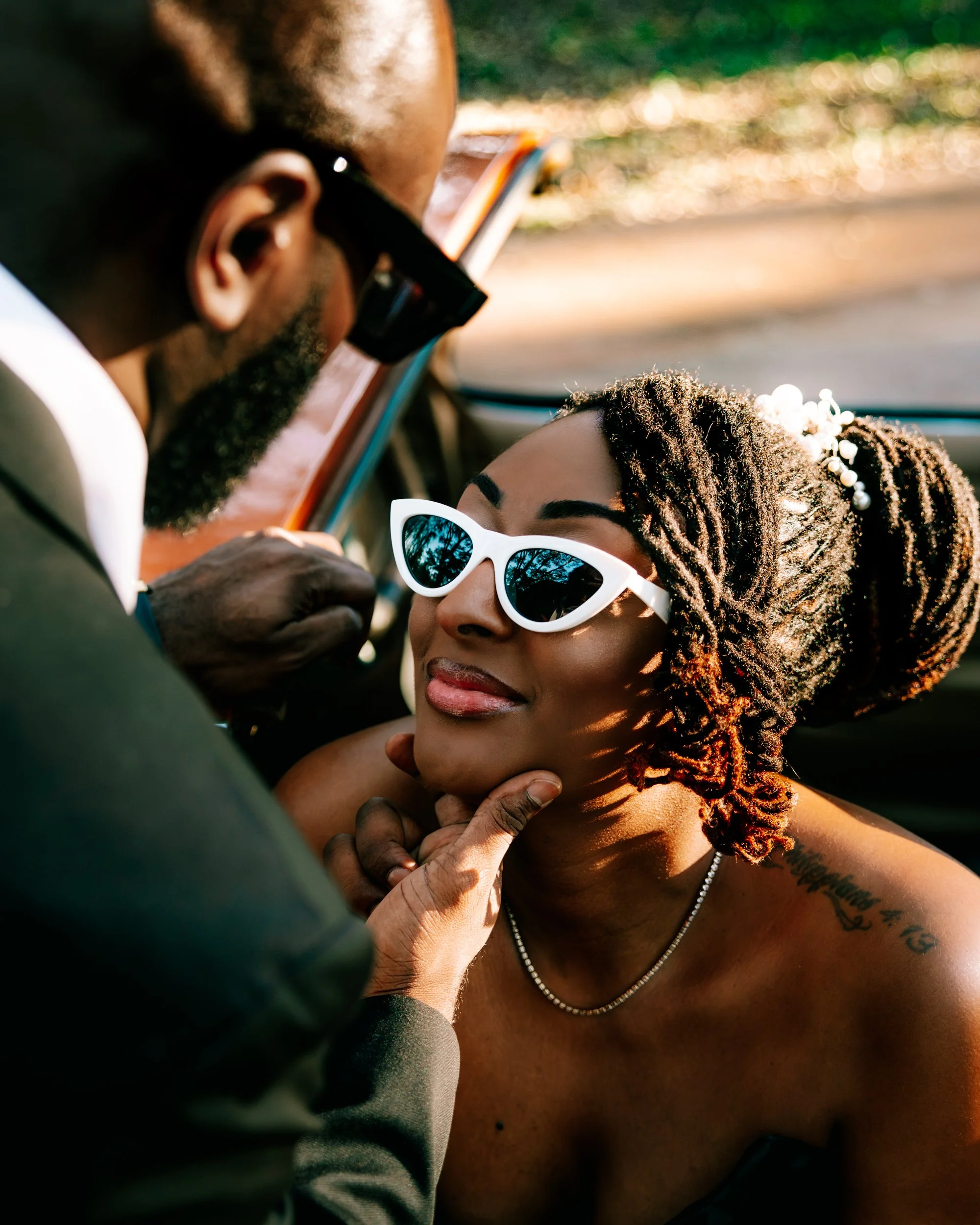 A man with glasses is holding a woman's chin, who is wearing white sunglasses and smiling softly. The woman has dreadlocks with beads and a tattoo on her shoulder, sitting inside a car.