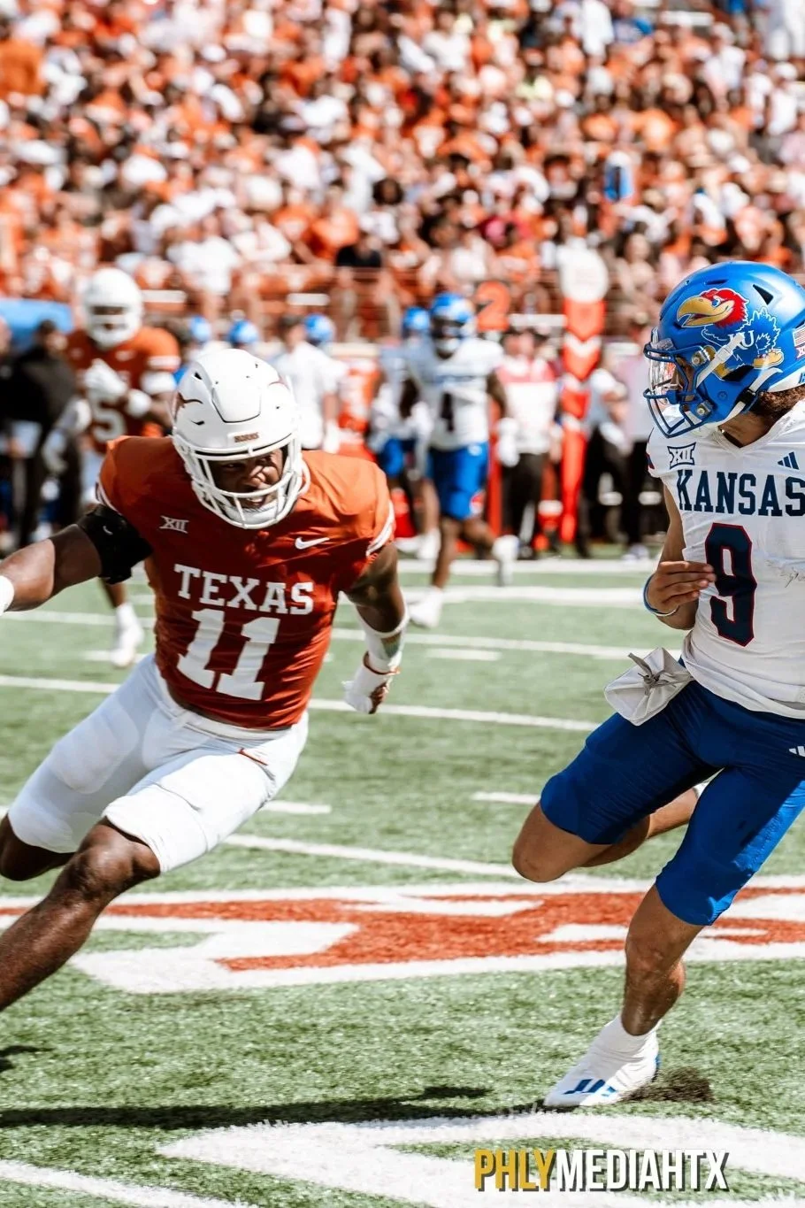 Two college football players in game action on the field, one from Texas wearing a red uniform with number 11, and the other from Kansas wearing a white uniform with number 9, chasing each other with a crowd in the stands in the background.