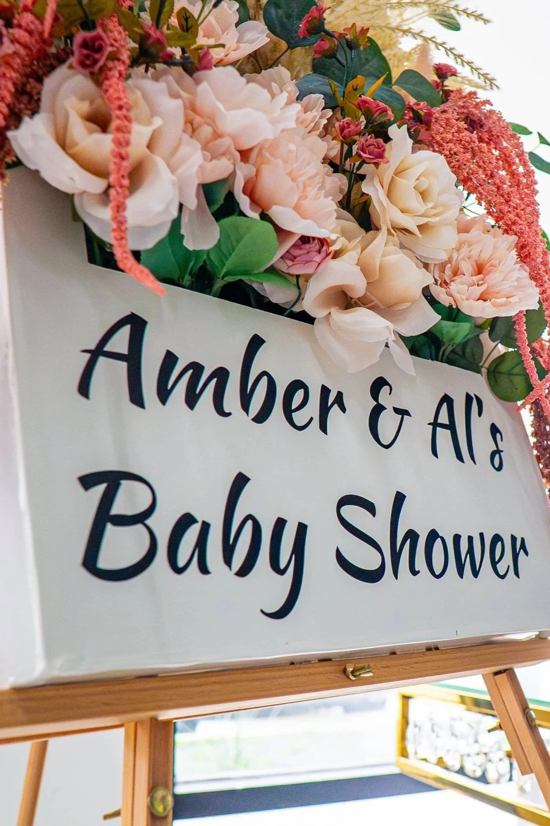 A white sign with black text reading "Amber & Ali's Baby Shower" surrounded by pink, peach, and cream-colored flowers and green leaves.