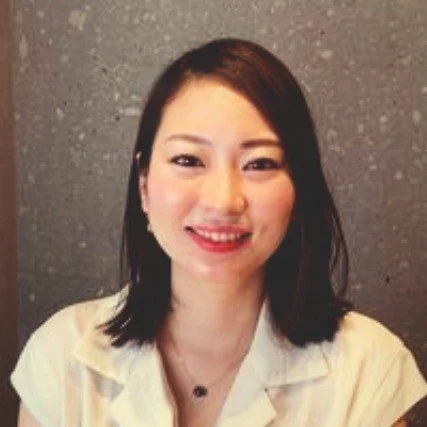 A young woman with dark hair smiling in front of a gray textured wall.