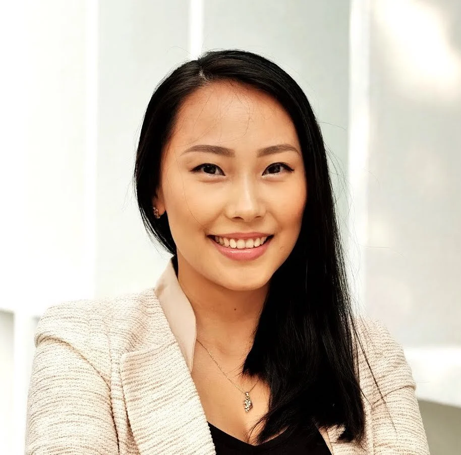 A young woman with long black hair smiling, wearing a beige blazer and a black top, in an indoor setting with blurred background.