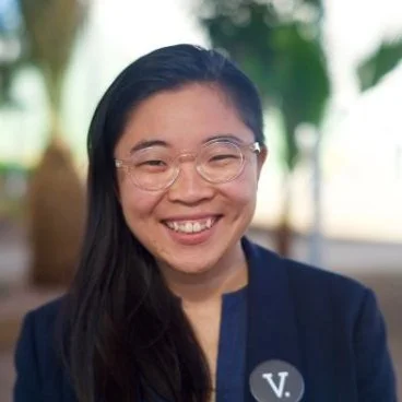 Woman smiling outdoors wearing glasses and a navy blue blazer with a circular badge marked 'V'.