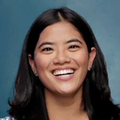 Smiling woman with dark hair, wearing earrings and a patterned top, against a blue background.