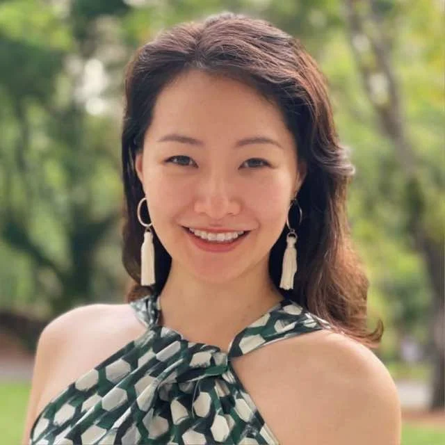 A woman with shoulder-length dark hair, smiling outdoors with green trees in the background, wearing large white earrings and a sleeveless top with a patterned scarf tied around her neck.