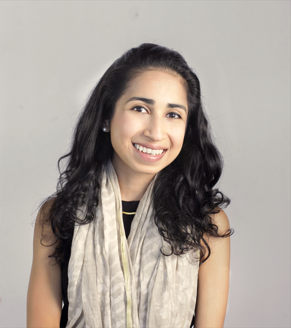 A young woman with long dark curly hair, smiling, wearing earrings, a black top with a light-colored scarf, and posing against a plain gray background.